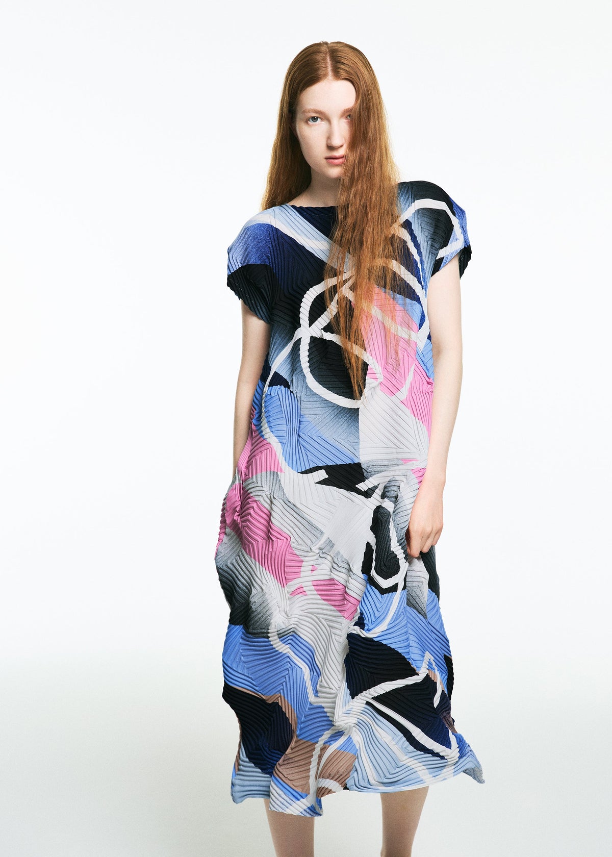 A woman with long red hair stands against a white background, wearing the TYPE-XV SOAR NY project 001 dress by A-POC ABLE ISSEY MIYAKE, featuring abstract blue, pink, white, and black patterns and embodying refined Japanese style. view 5 of 8