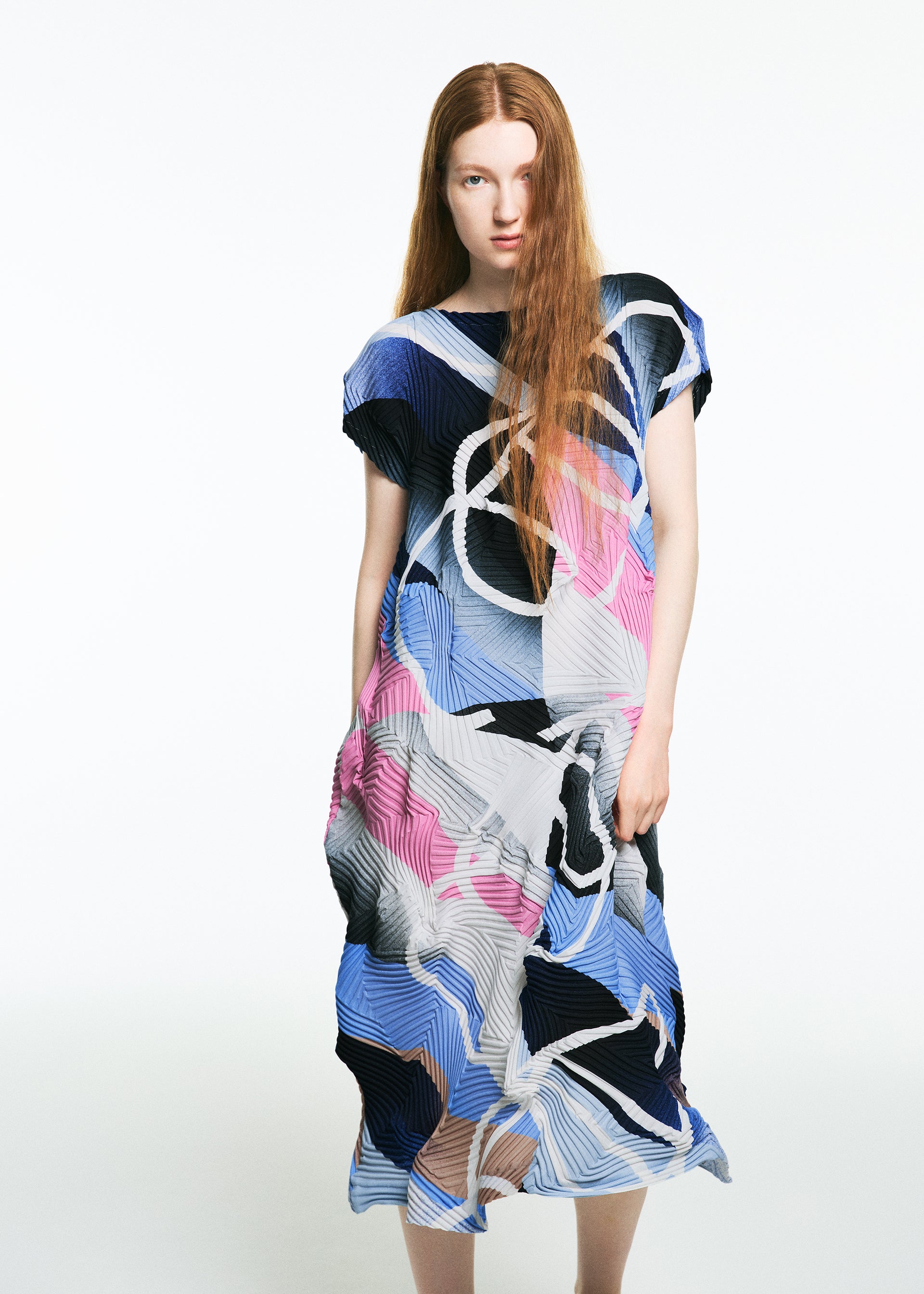 Zoomed view A woman with long red hair stands against a white background, wearing the TYPE-XV SOAR NY project 001 dress by A-POC ABLE ISSEY MIYAKE, featuring abstract blue, pink, white, and black patterns and embodying refined Japanese style.