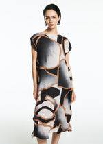 A person with short dark hair wears the TYPE-XV SOAR NY project 001 dress by A-POC ABLE ISSEY MIYAKE, featuring an abstract geometric pattern in black, white, beige, and peach, standing against a plain white background.