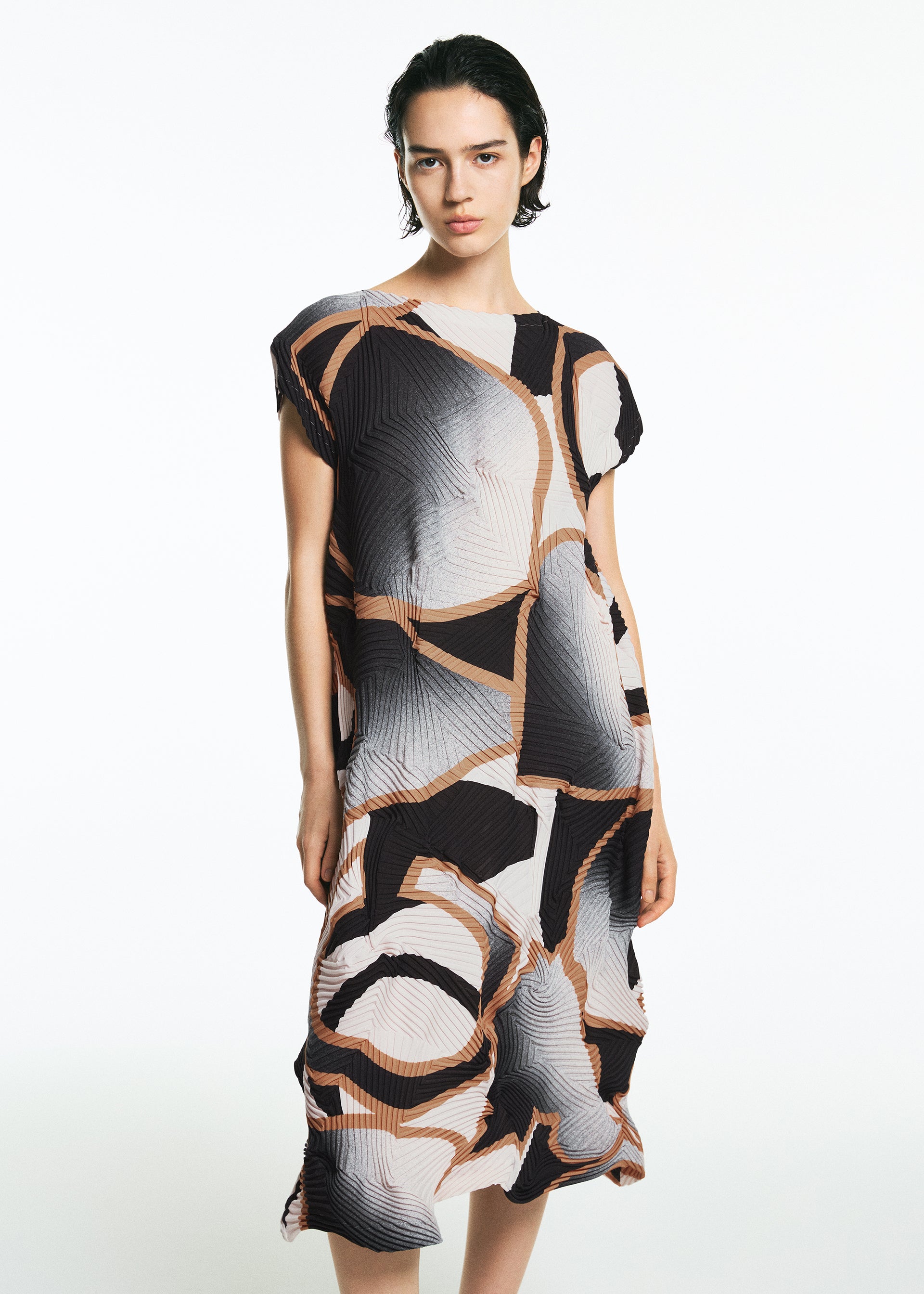 Zoomed view A person with short dark hair wears the TYPE-XV SOAR NY project 001 dress by A-POC ABLE ISSEY MIYAKE, featuring an abstract geometric pattern in black, white, beige, and peach, standing against a plain white background.