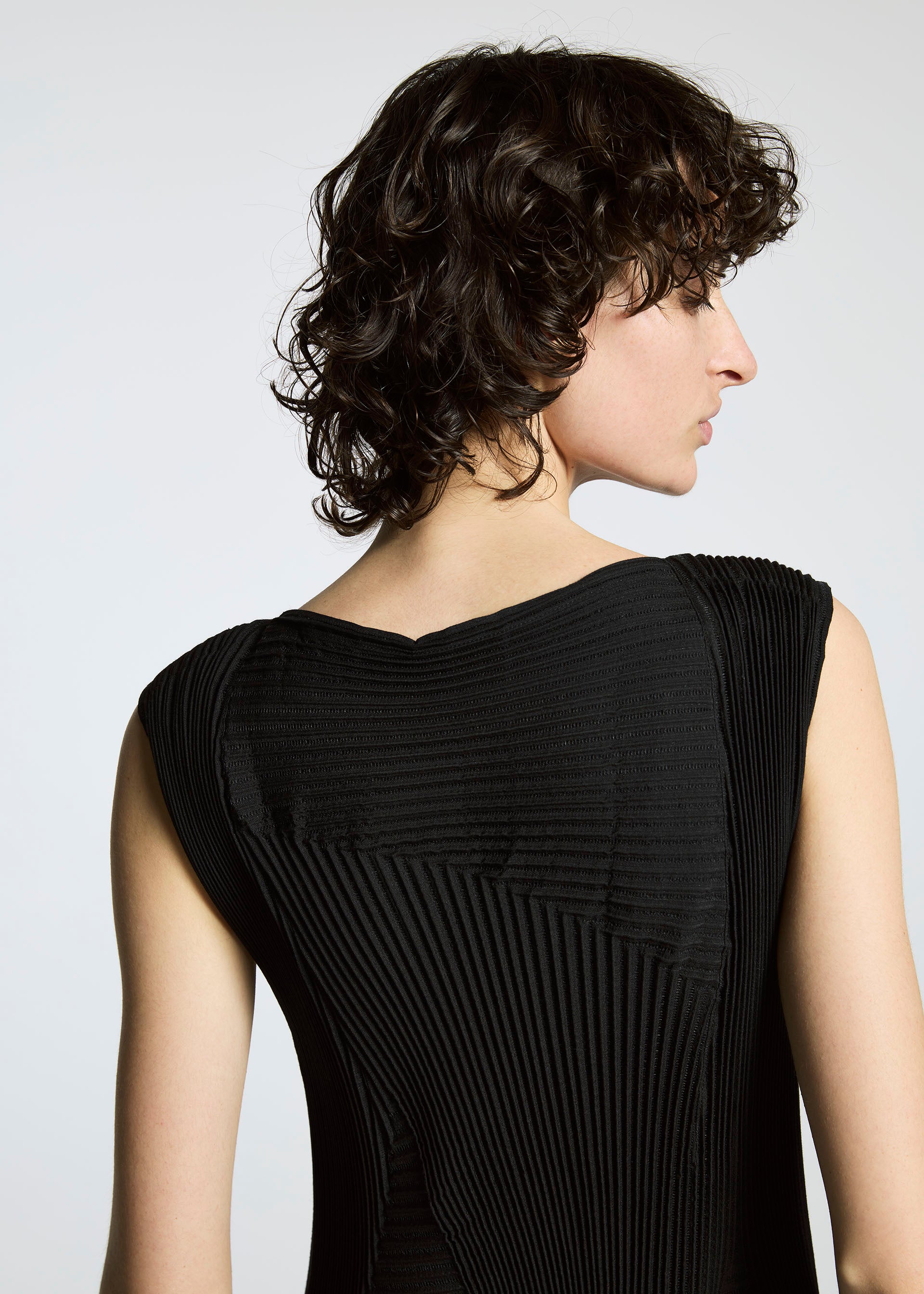 Zoomed view A person with short, curly dark hair, viewed from behind and in slight profile, wears a black textured A-POC ABLE ISSEY MIYAKE TYPE-Ⅰ 008 dress against a neutral background.