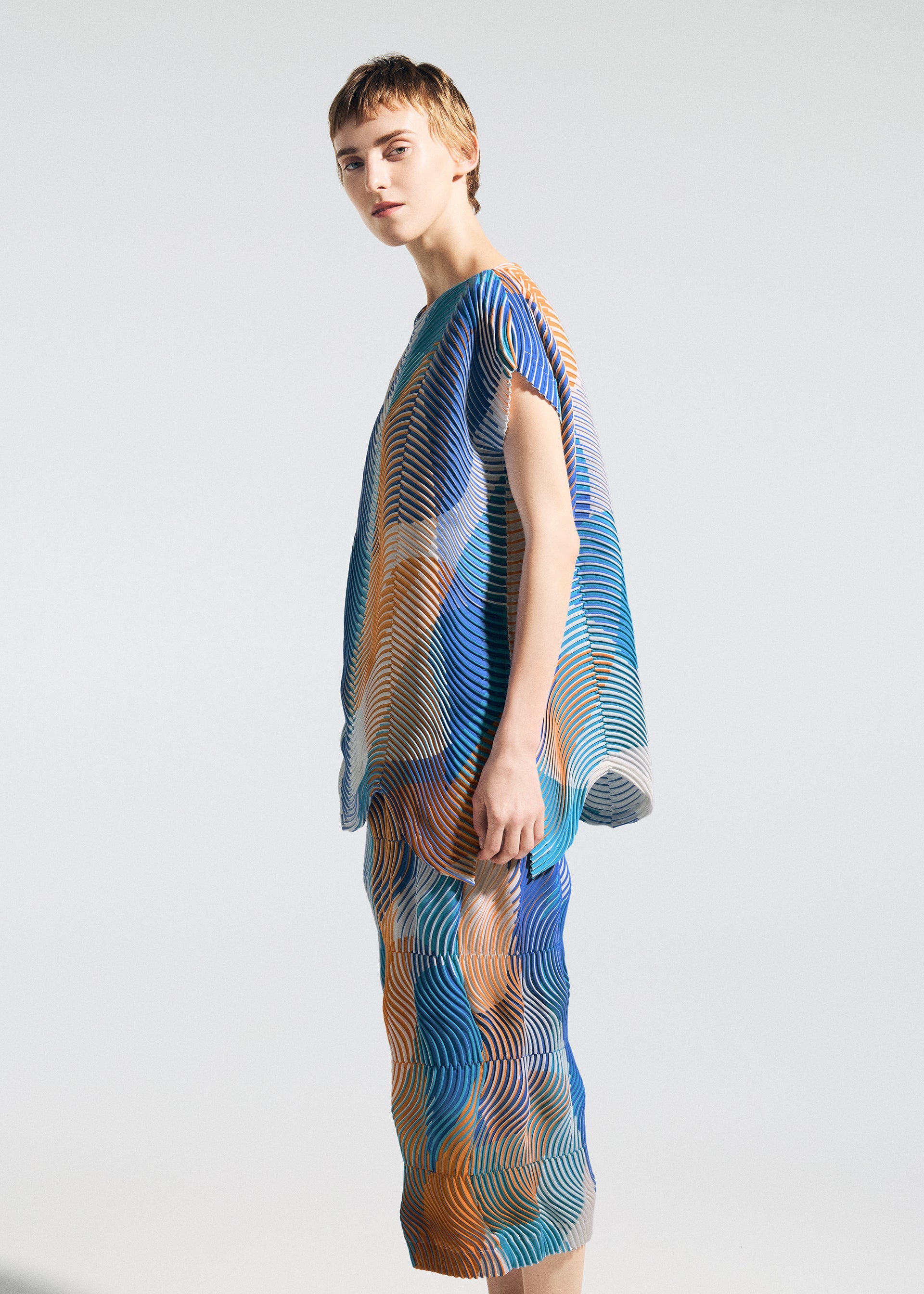 Zoomed view A person stands sideways in the TYPE-P 005 by A-POC ABLE ISSEY MIYAKE, featuring a loose outfit with wavy blue, orange, and white patterns on a French-sleeved top and pants, set against a plain light background.