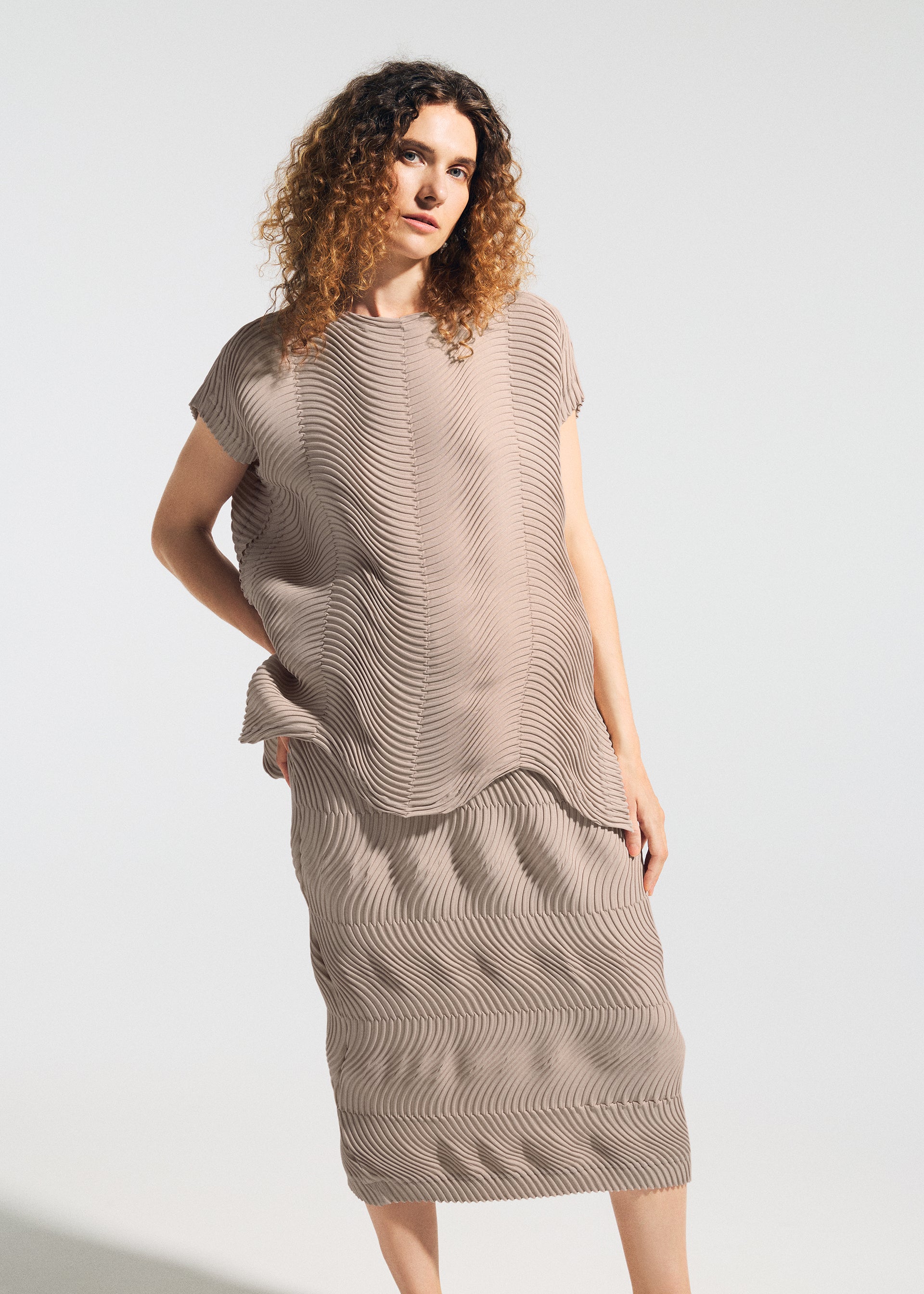 Zoomed view A woman with curly hair wears the TYPE-P 006 French-sleeved top and matching skirt by A-POC ABLE ISSEY MIYAKE, featuring a taupe wavy texture—versatile for everyday wear—standing against a plain light background.