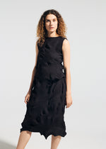 A woman with curly hair wears the TYPE-P 003-2, a sleeveless black dress by A-POC ABLE ISSEY MIYAKE, featuring a unique wavy pattern and Baked Stretch texture. She stands against a plain light background, looking to the side with a neutral expression.