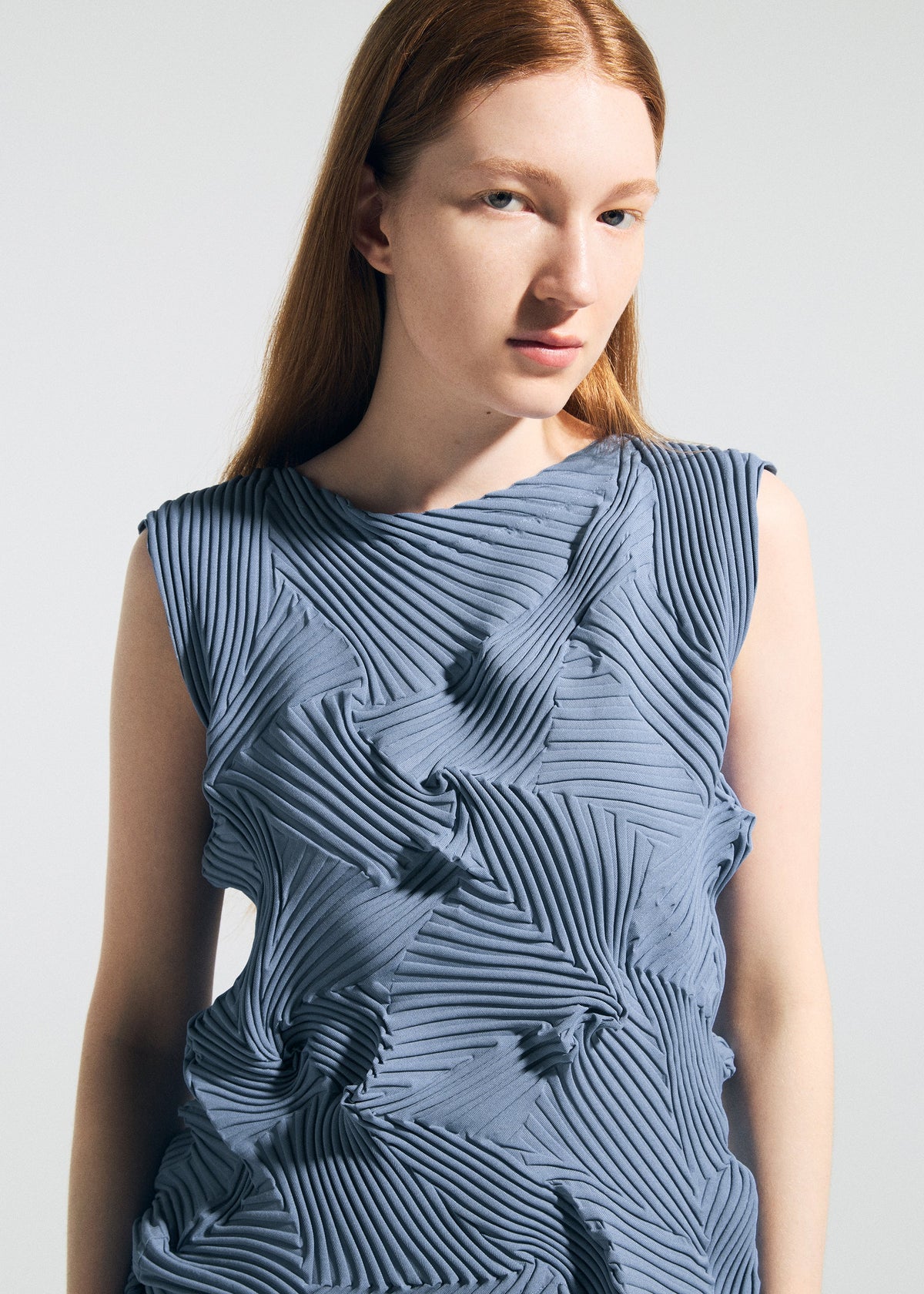 A woman with long red hair wears the TYPE-P 003-2 sleeveless blue top by A-POC ABLE ISSEY MIYAKE, featuring geometric pleated patterns made with the Baked Stretch technique, as she gazes to the side against a plain light background. view 8 of 10