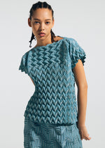 A person with braided hair wears the TYPE-O 009 set by A-POC ABLE ISSEY MIYAKE—a teal, short-sleeved top and matching pleated skirt—posed against a plain light background.