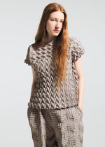 A woman with long red hair models the TYPE-O 009 short-sleeve top and pants by A-POC ABLE ISSEY MIYAKE, featuring geometric pleats and an origami-like texture. She stands with one hand in her pocket against a plain background.