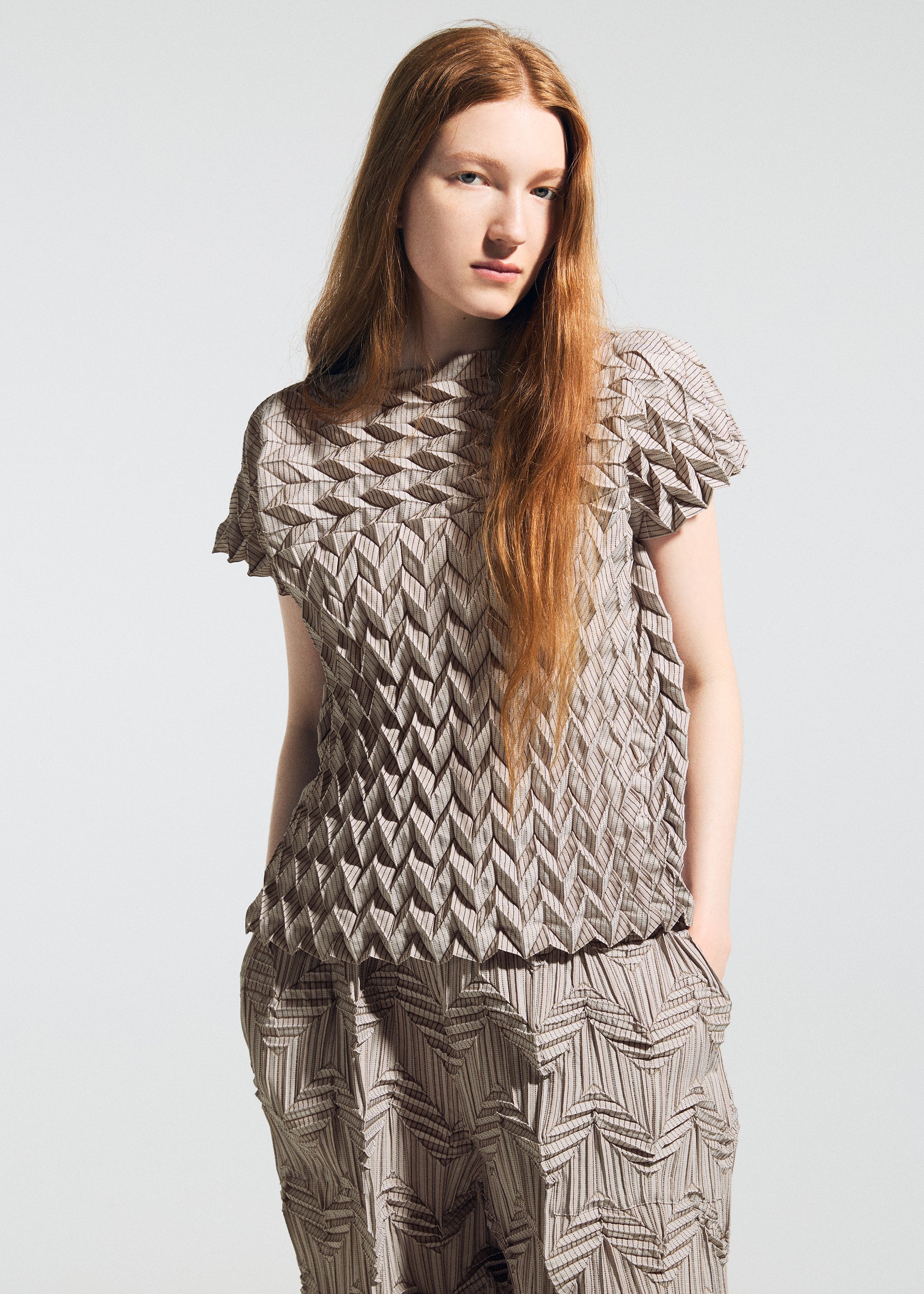 Zoomed view A woman with long red hair models the TYPE-O 009 short-sleeve top and pants by A-POC ABLE ISSEY MIYAKE, featuring geometric pleats and an origami-like texture. She stands with one hand in her pocket against a plain background.
