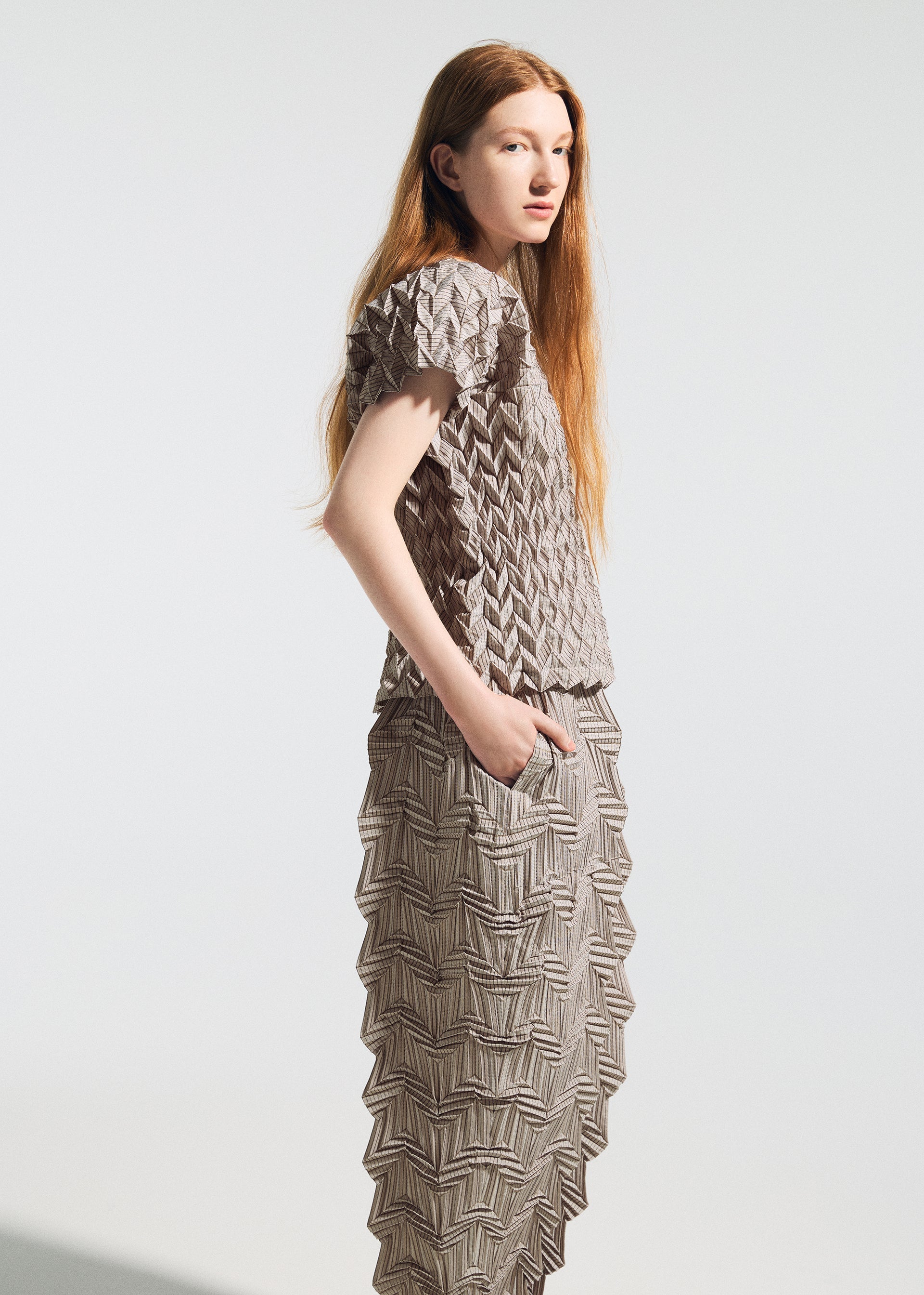 Zoomed view A person with long red hair models the TYPE-O 009 matching top and skirt by A-POC ABLE ISSEY MIYAKE, featuring textured geometric zigzag pleats and a unique shape, standing with one hand in a pocket against a plain light background.