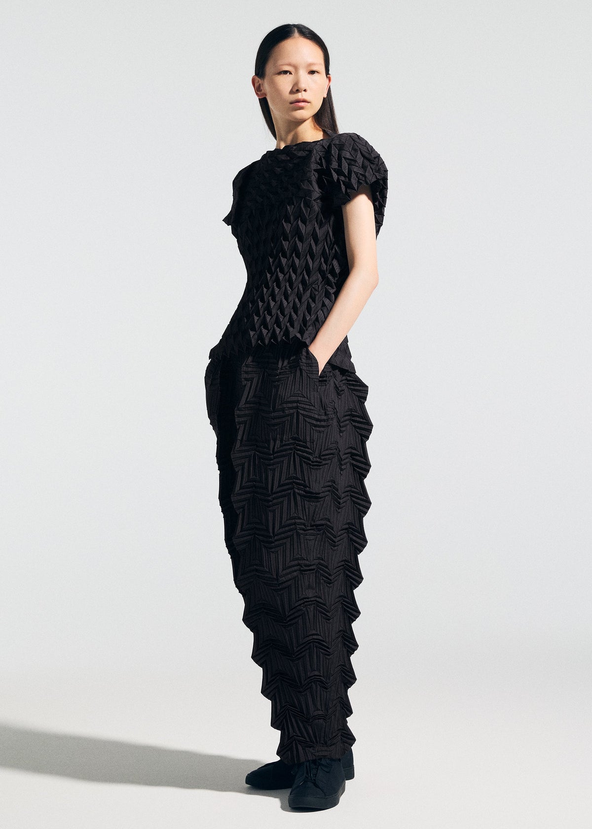 A woman with long black hair stands against a plain light background, wearing the TYPE-O 009 set by A-POC ABLE ISSEY MIYAKE—black textured top and pants with geometric pleats and 3D patterns—hands in pockets, paired with black shoes. view 8 of 9