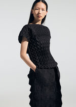 A woman with straight dark hair models the TYPE-O 009 short-sleeve black geometric pleated top and skirt by A-POC ABLE ISSEY MIYAKE, standing with one hand in her pocket against a plain light background.