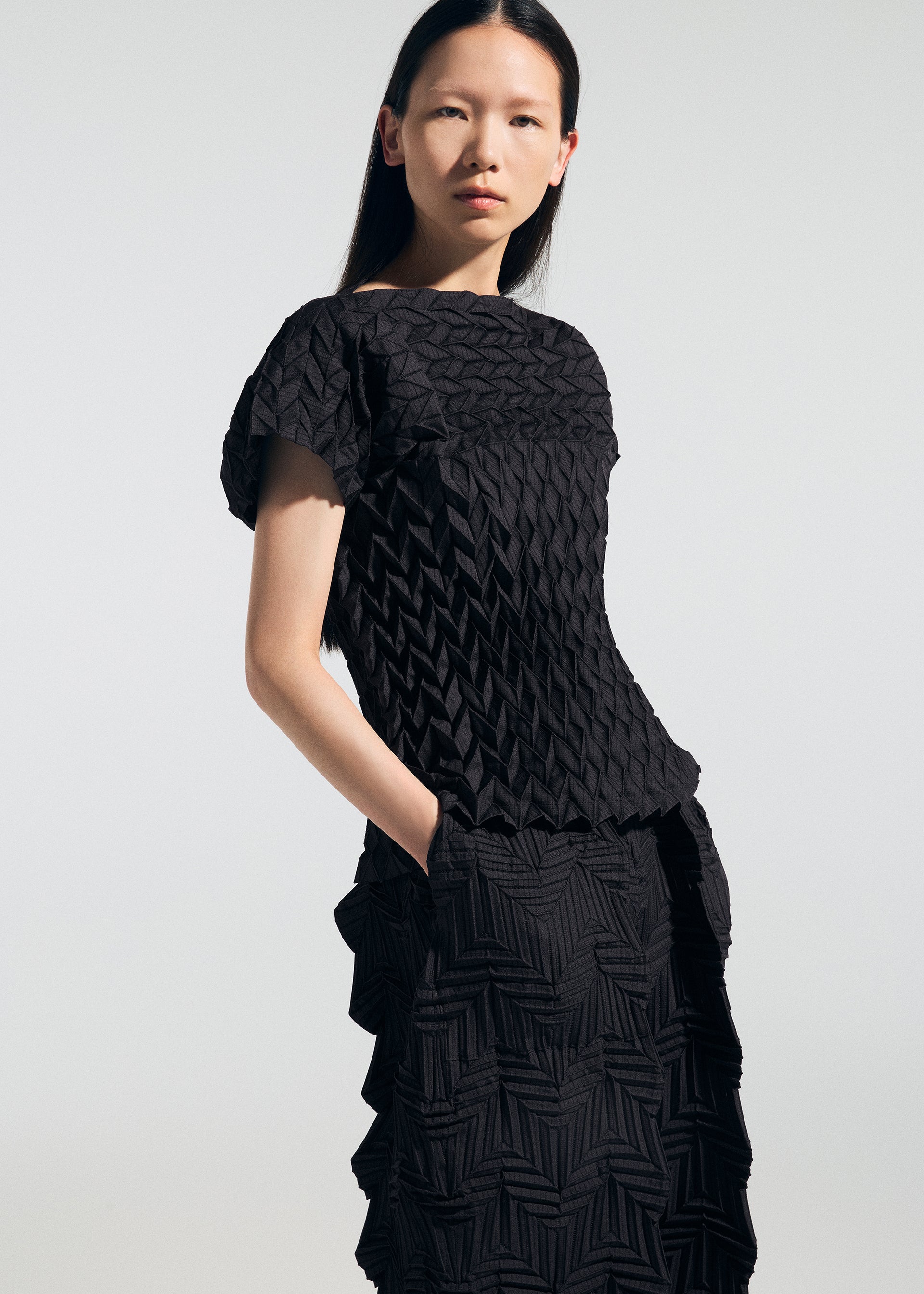 Zoomed view A woman with straight dark hair models the TYPE-O 009 short-sleeve black geometric pleated top and skirt by A-POC ABLE ISSEY MIYAKE, standing with one hand in her pocket against a plain light background.