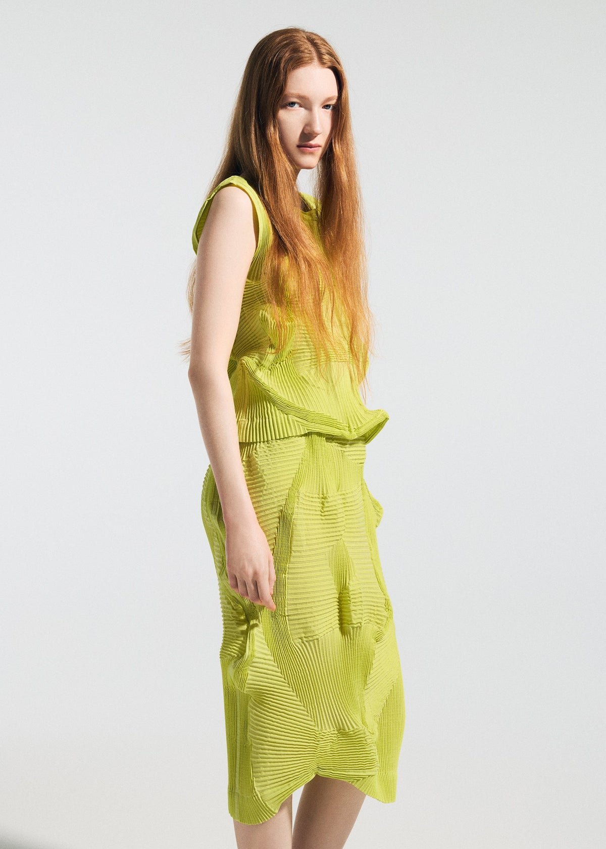 A woman with long red hair stands against a plain background, wearing the TYPE-O 022 dress by A-POC ABLE ISSEY MIYAKE in yellow-green moisture-regulating fabric. She gazes to the side with a neutral expression. view 5 of 6