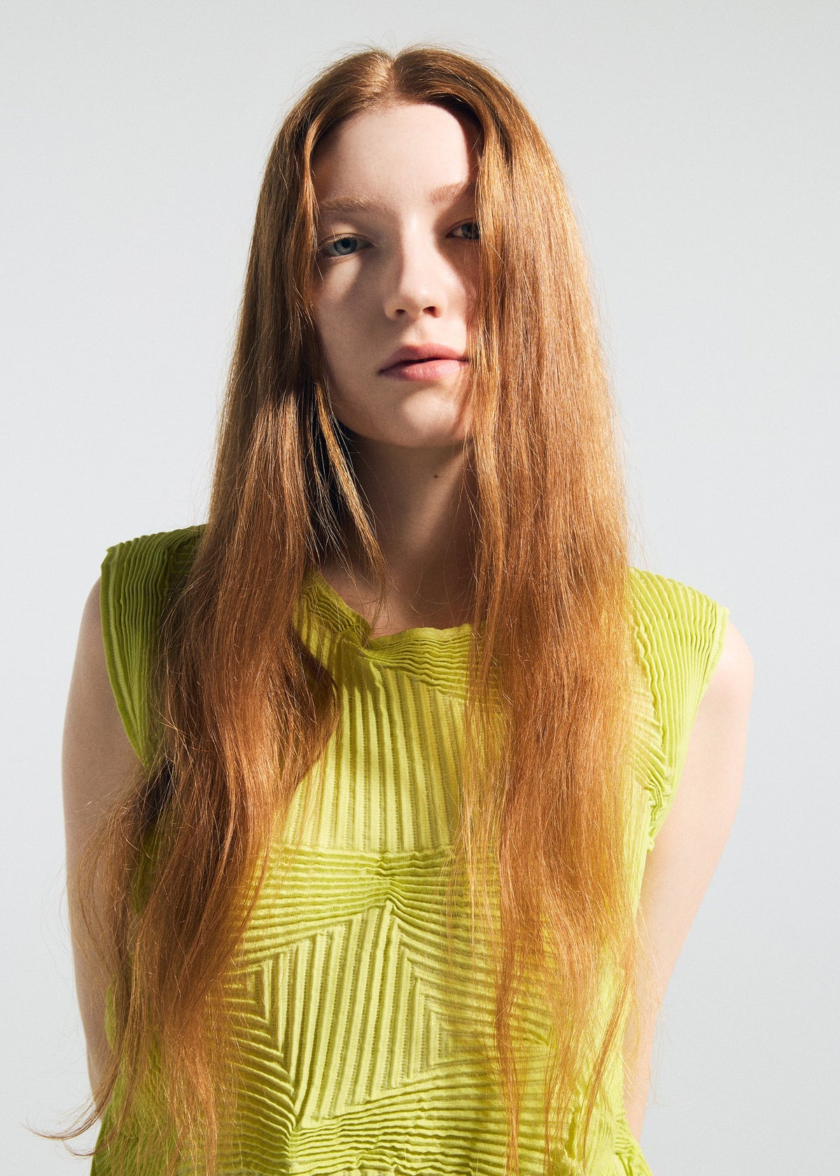 A young woman with long, straight red hair wears the TYPE-O 022 antibacterial sleeveless top by A-POC ABLE ISSEY MIYAKE in bright yellow-green, standing against a plain light background and looking at the camera with a neutral expression. view 6 of 6