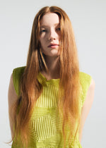 A young woman with long, straight red hair wears the TYPE-O 022 antibacterial sleeveless top by A-POC ABLE ISSEY MIYAKE in bright yellow-green, standing against a plain light background and looking at the camera with a neutral expression.