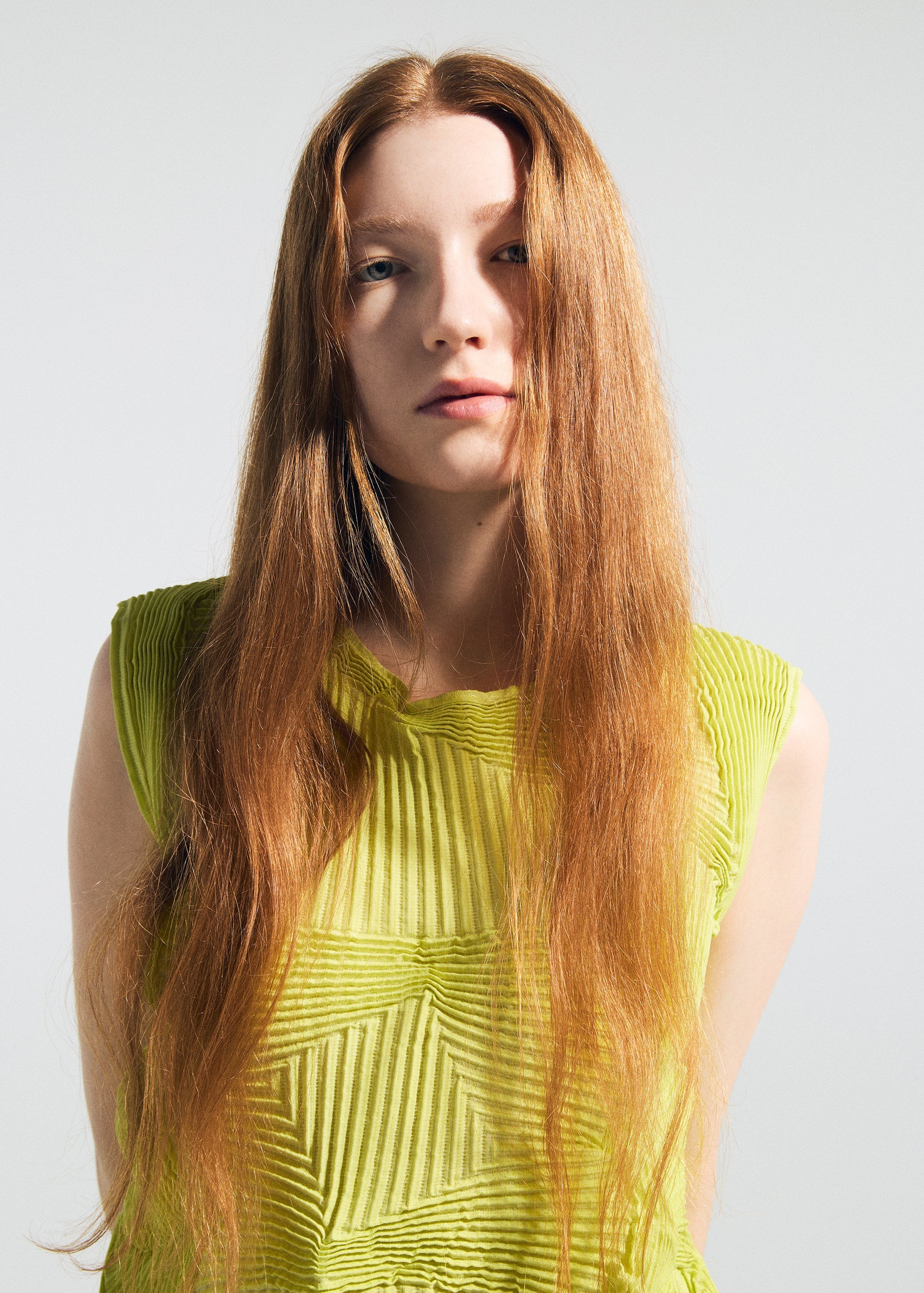 Zoomed view A young woman with long, straight red hair wears the TYPE-O 022 antibacterial sleeveless top by A-POC ABLE ISSEY MIYAKE in bright yellow-green, standing against a plain light background and looking at the camera with a neutral expression.