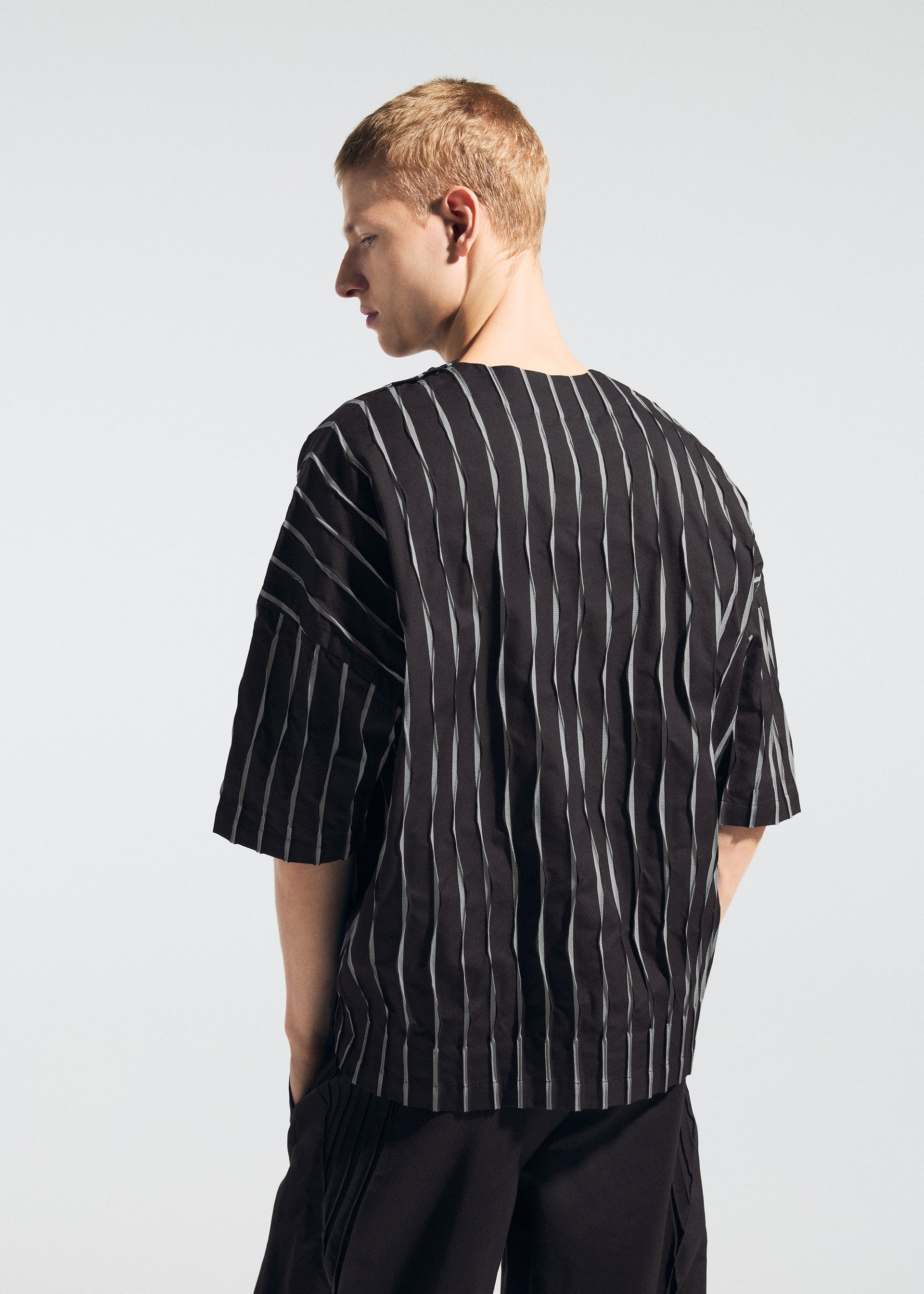Zoomed view A person with short blond hair stands sideways, looking over their shoulder, wearing the TYPE-W 011 top by A-POC ABLE ISSEY MIYAKE— a loose black piece with vertical white stripes and paired with black pants against a light background.