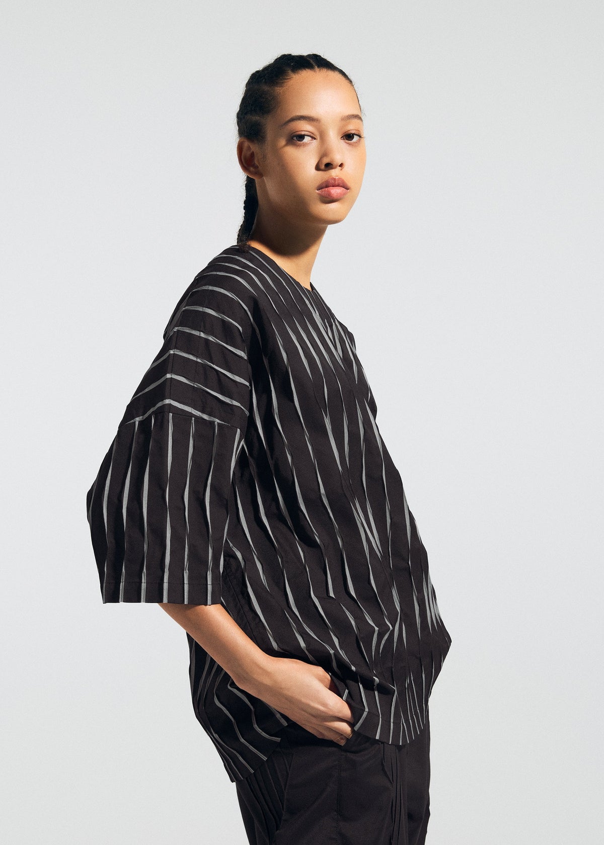 A person with braided hair poses relaxedly in the TYPE-W 011 set by A-POC ABLE ISSEY MIYAKE: a loose black top with white stripes and black pants with zigzag pleats, against a plain light gray background. view 7 of 10