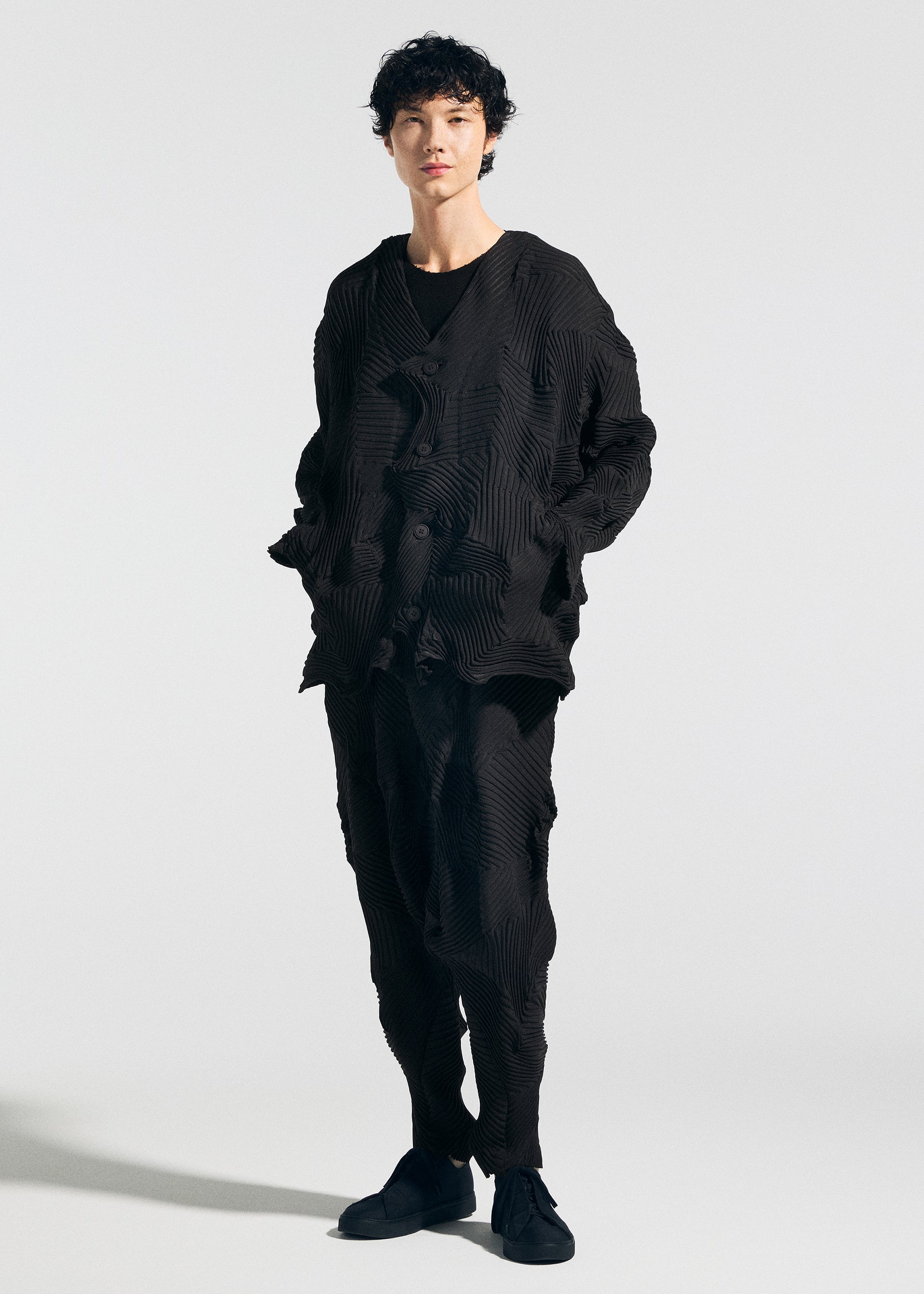 Zoomed view A person stands against a plain light background wearing the TYPE-P 003-1 jacket and pants by A-POC ABLE ISSEY MIYAKE, featuring Baked Stretch pleating and a soft texture. They pair the look with black shoes, hands in pockets, facing the camera.