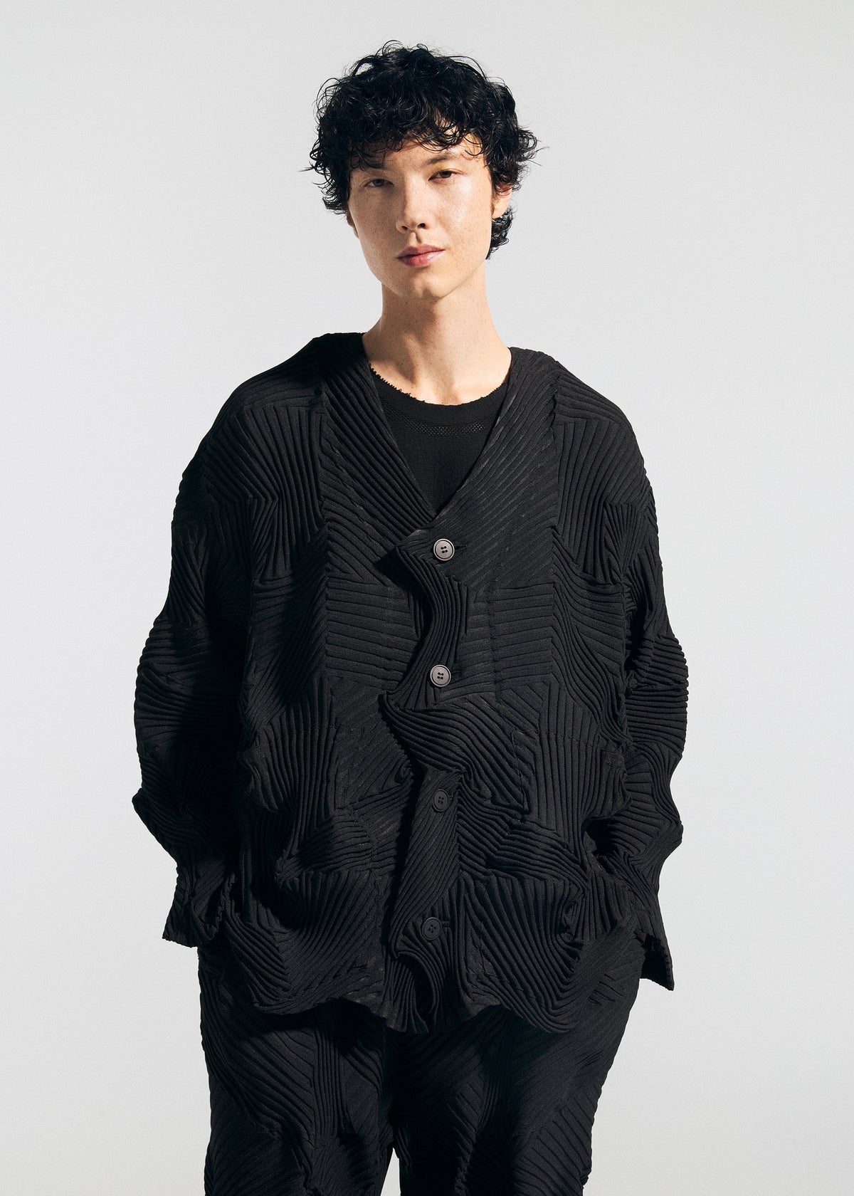 Person with short, curly dark hair wearing the TYPE-P 003-1 textured black jacket and matching pants by A-POC ABLE ISSEY MIYAKE, hands in pockets, standing against a plain light background. view 7 of 8