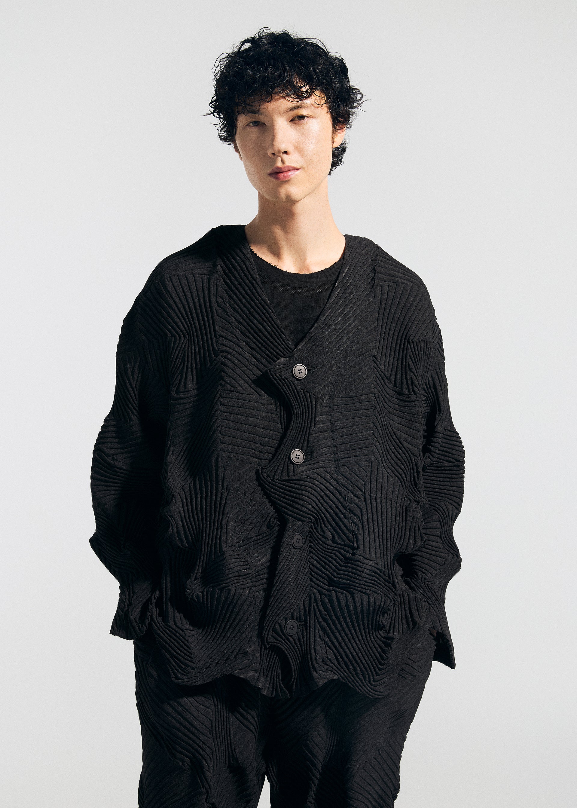 Zoomed view Person with short, curly dark hair wearing the TYPE-P 003-1 textured black jacket and matching pants by A-POC ABLE ISSEY MIYAKE, hands in pockets, standing against a plain light background.