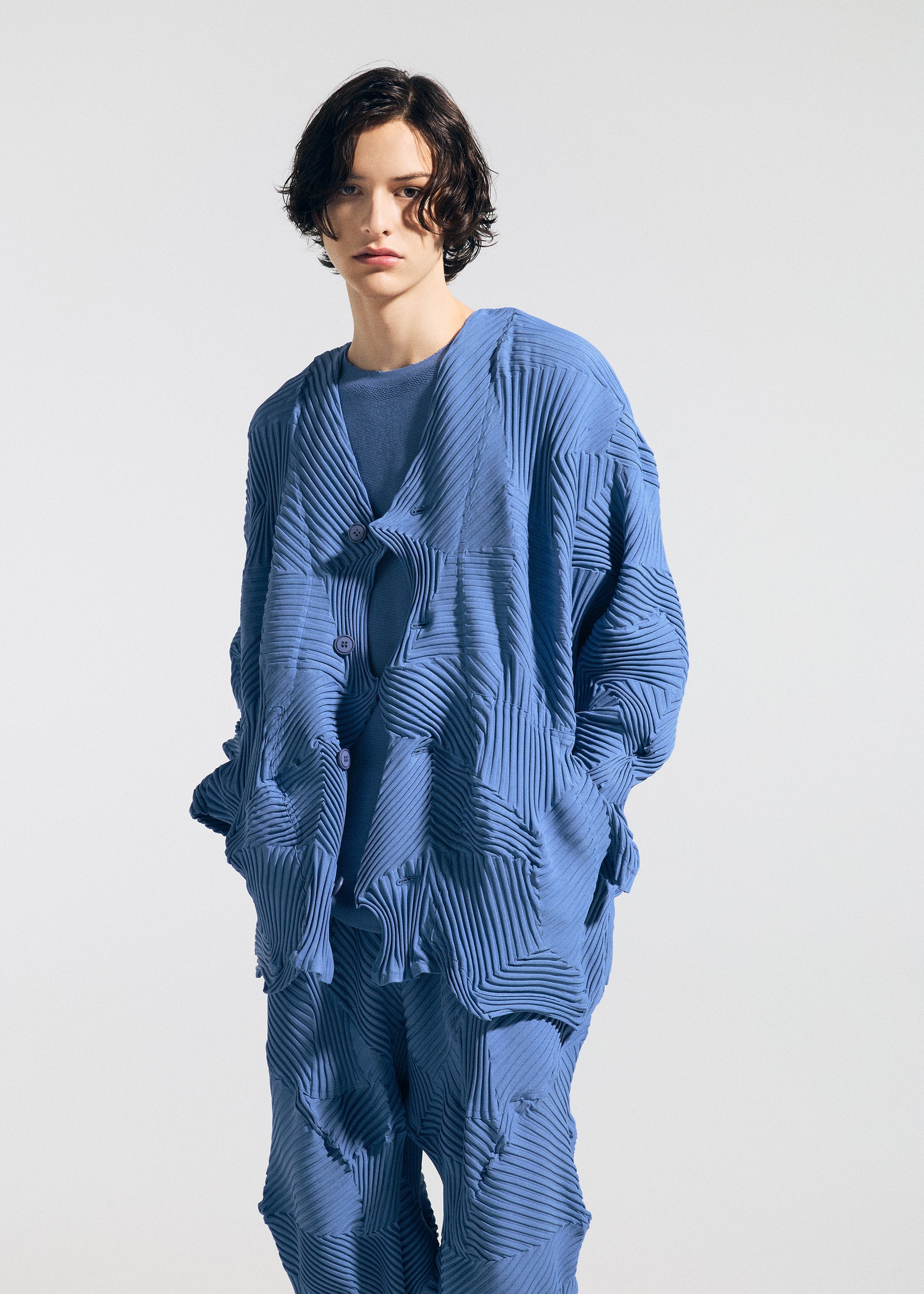 Zoomed view A person with short, wavy dark hair wears the TYPE-P 003-1 by A-POC ABLE ISSEY MIYAKE, a loose blue outfit with a patterned jacket and matching pants, showcasing soft texture and Baked Stretch pleating against a plain light background.