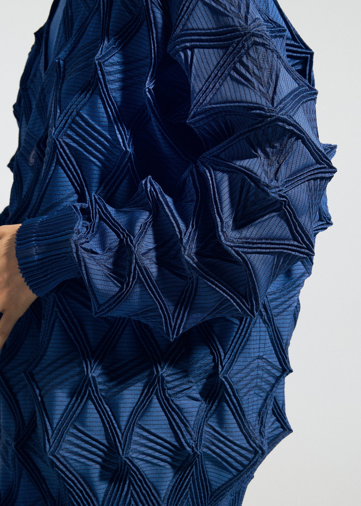 A close-up of a person wearing TYPE-O 023 by A-POC ABLE ISSEY MIYAKE, a blue textured garment featuring raised, geometric, origami-like patterns that create a striking three-dimensional effect. The fabric showcases intricate, angular folds and lines reminiscent of Steam Stretch techniques throughout. view 7 of 11