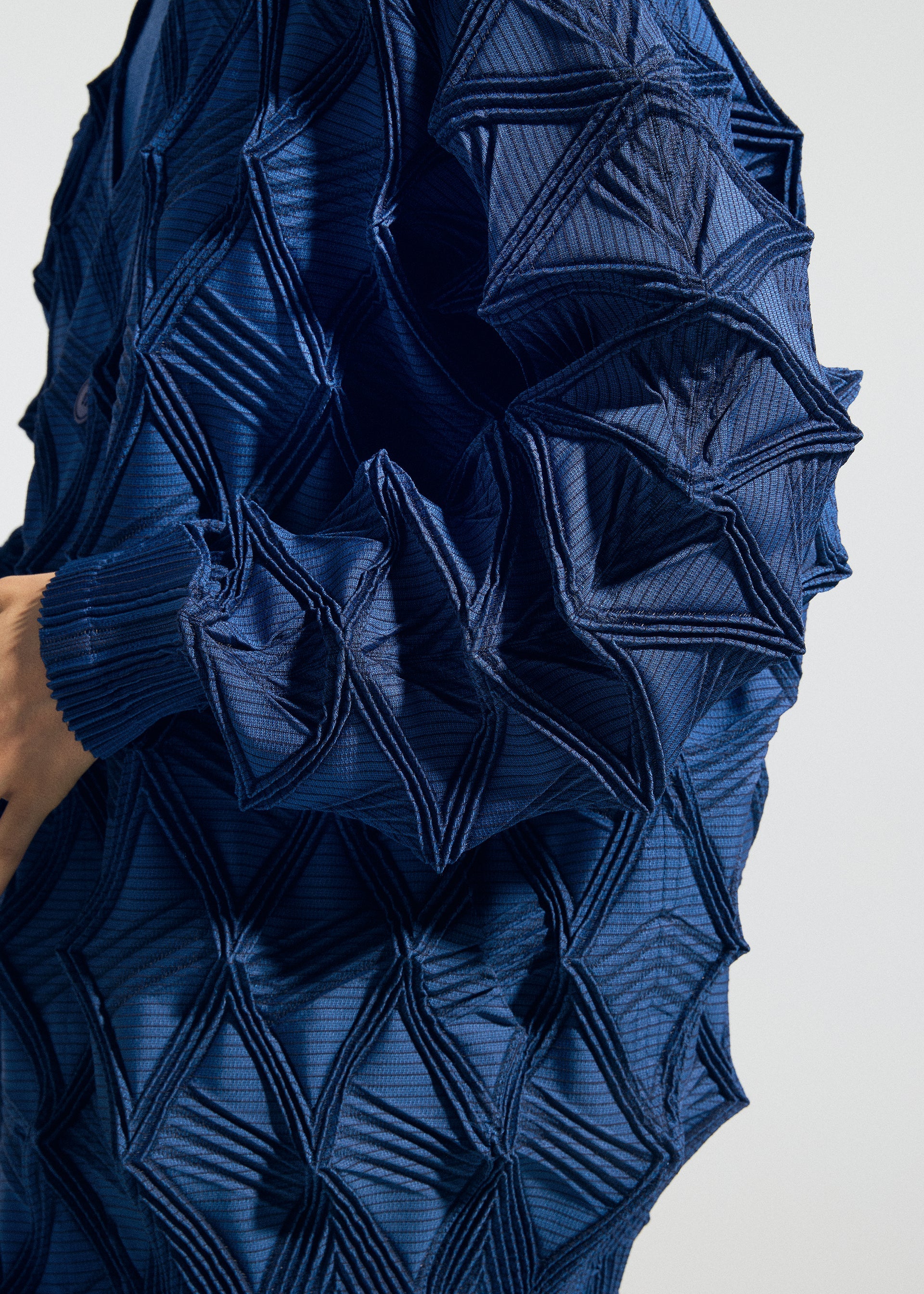 Zoomed view A close-up of a person wearing TYPE-O 023 by A-POC ABLE ISSEY MIYAKE, a blue textured garment featuring raised, geometric, origami-like patterns that create a striking three-dimensional effect. The fabric showcases intricate, angular folds and lines reminiscent of Steam Stretch techniques throughout.