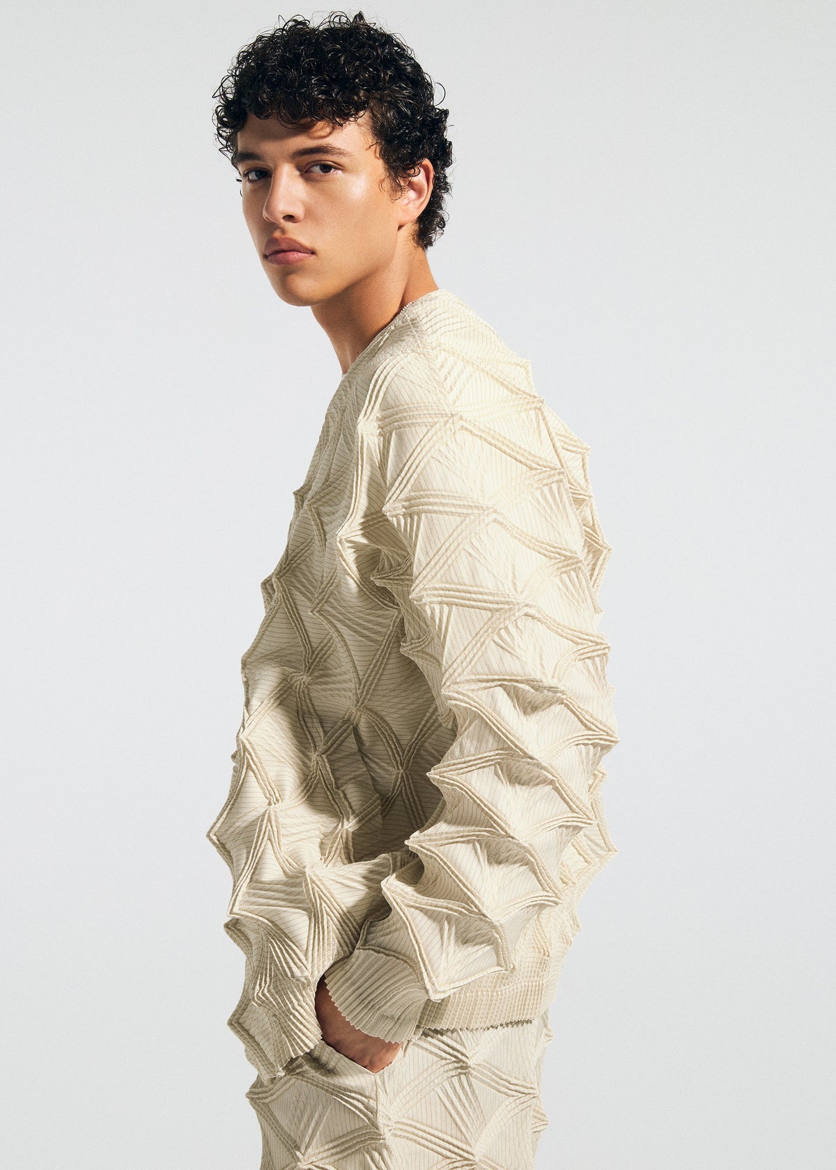 A young person with short curly hair stands in profile against a plain white background, wearing the TYPE-O 023 textured cream-colored sweater with raised geometric patterns and pleated pants from A-POC ABLE ISSEY MIYAKE. view 9 of 11