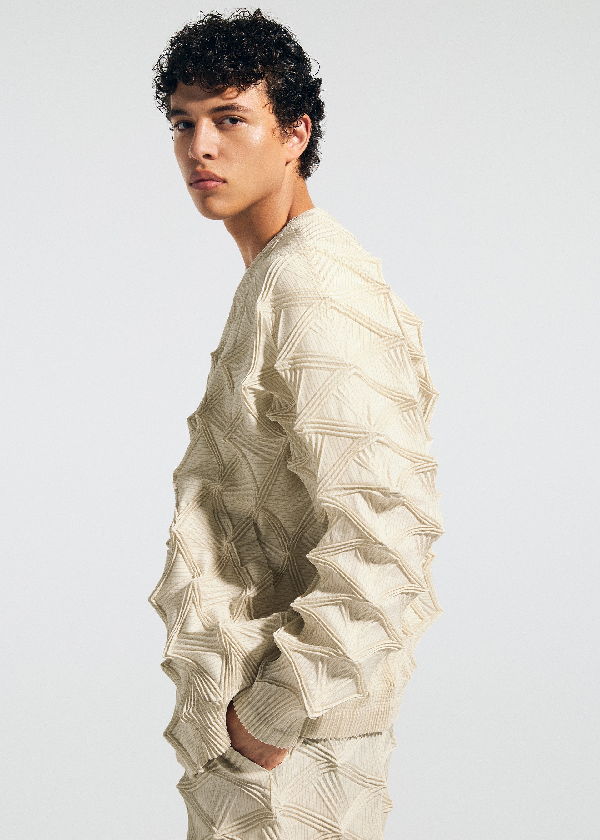 Zoomed view A young person with short curly hair stands in profile against a plain white background, wearing the TYPE-O 023 textured cream-colored sweater with raised geometric patterns and pleated pants from A-POC ABLE ISSEY MIYAKE.