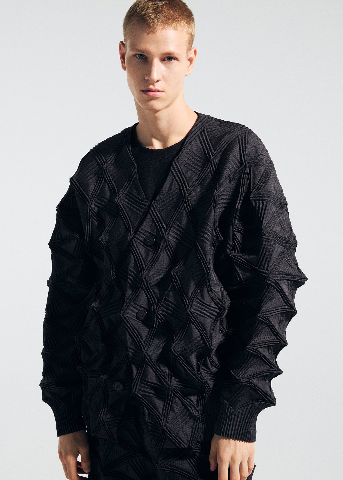 A person wears the TYPE-O 023 sweater by A-POC ABLE ISSEY MIYAKE—featuring a textured black geometric pattern—and pairs it with pleated pants, standing against a plain light background while looking directly at the camera with a neutral expression. view 11 of 11