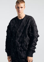 A person wears the TYPE-O 023 sweater by A-POC ABLE ISSEY MIYAKE—featuring a textured black geometric pattern—and pairs it with pleated pants, standing against a plain light background while looking directly at the camera with a neutral expression.