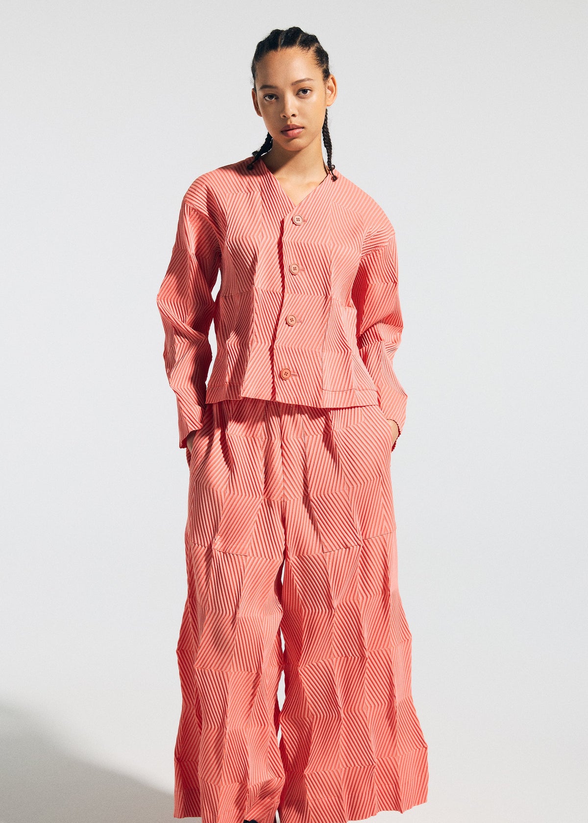 A person with braided hair models the TYPE-W 009-1 matching set by A-POC ABLE ISSEY MIYAKE: a relaxed fit, long-sleeve button-up top and wide-leg pants in pink and white geometric stripes, crafted from stretchy polyester, against a plain background. view 6 of 6