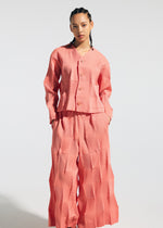A person with braided hair models the TYPE-W 009-1 matching set by A-POC ABLE ISSEY MIYAKE: a relaxed fit, long-sleeve button-up top and wide-leg pants in pink and white geometric stripes, crafted from stretchy polyester, against a plain background.