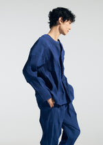 A person with short curly hair wears the TYPE-W 009-2 cardigan and matching pants by A-POC ABLE ISSEY MIYAKE, crafted in blue textured stretch polyester. Standing in profile with hands in pockets against a plain light background, the relaxed-fit ensemble offers a comfortable yet modern look.