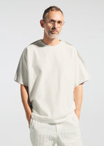 A man with short hair, glasses, and a beard stands against a light background, wearing the loose-fitting TYPE-J 001 top and pants by A-POC ABLE ISSEY MIYAKE, both off-white with frayed edges and his hands in his pockets.