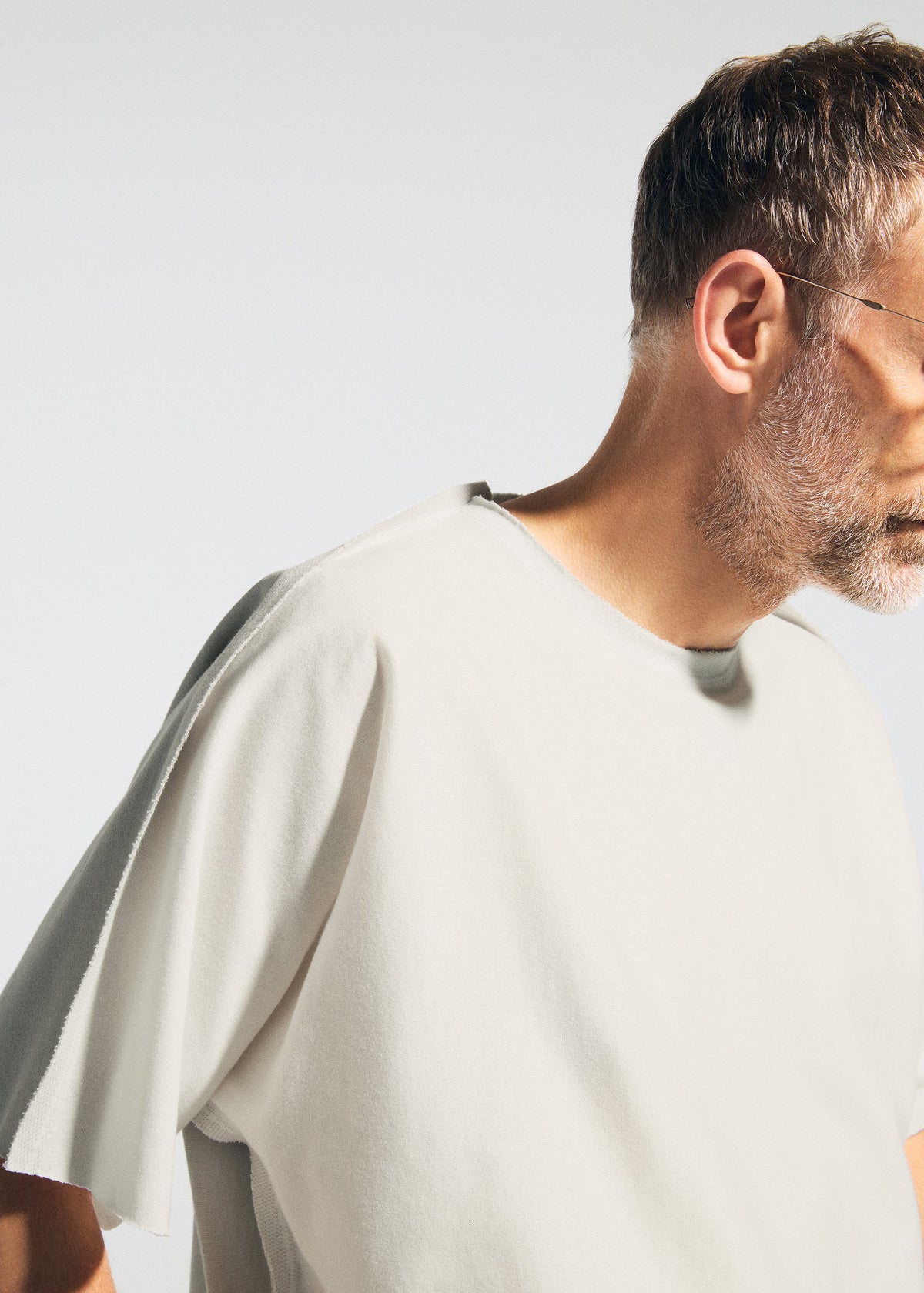 A man with short hair, a beard, and glasses wears the TYPE-J 001 two-tone knit oversized t-shirt by A-POC ABLE ISSEY MIYAKE, shown from the side against a light background with his face partially out of frame. view 7 of 11