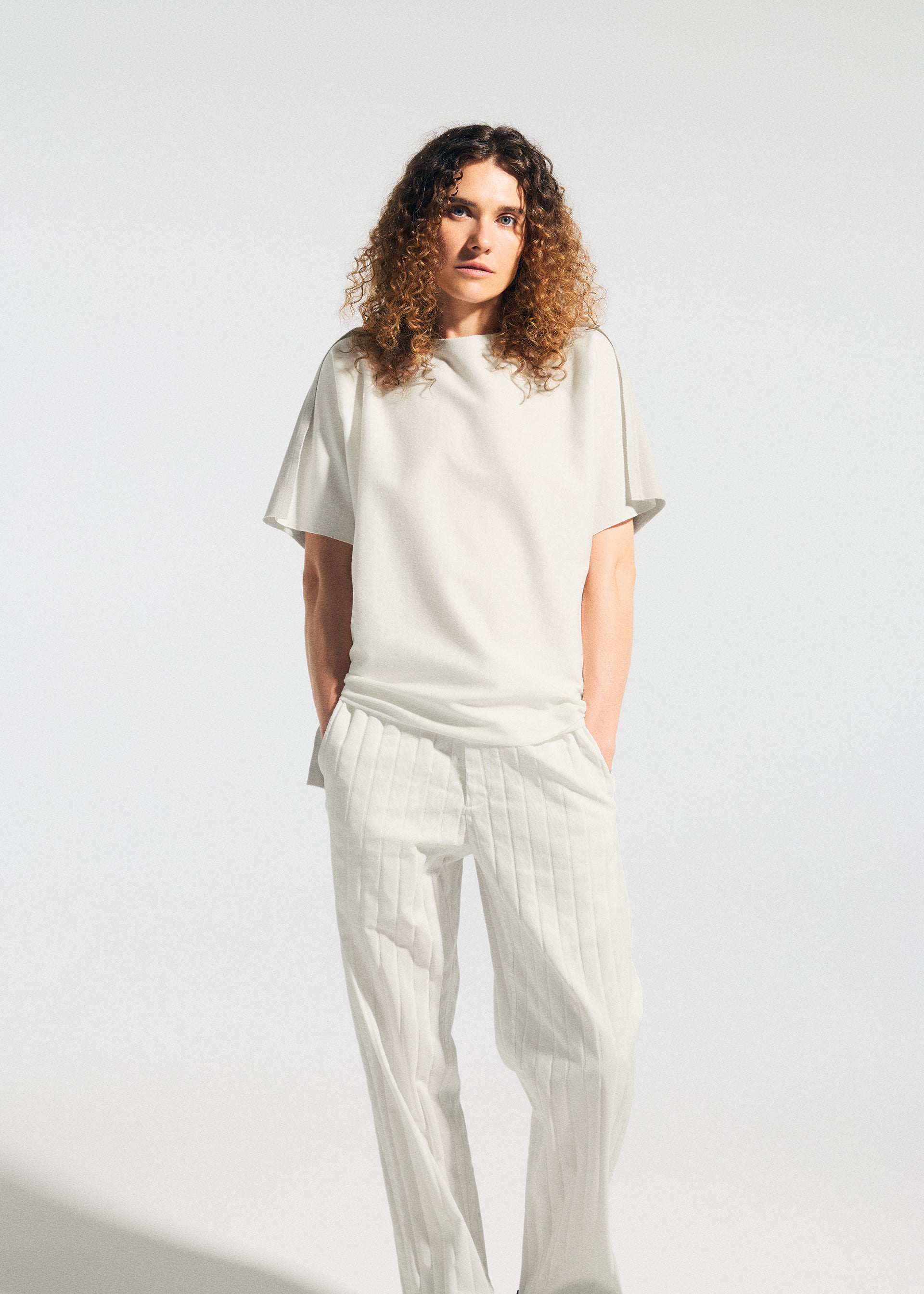 Zoomed view A person with curly hair stands against a plain background, wearing the TYPE-J 001 by A-POC ABLE ISSEY MIYAKE—a white frayed edge top and white pleated pants—with hands in their pockets.