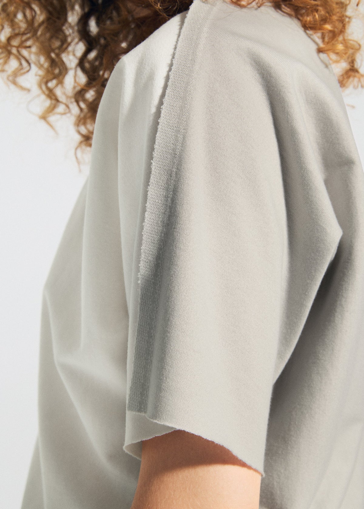 A close-up of a person wearing the TYPE-J 001 light gray top by A-POC ABLE ISSEY MIYAKE, featuring frayed edges and visible raw seams; the person's curly hair is partially visible. view 9 of 11