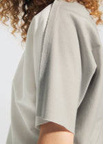 A close-up of a person wearing the TYPE-J 001 light gray top by A-POC ABLE ISSEY MIYAKE, featuring frayed edges and visible raw seams; the person's curly hair is partially visible.