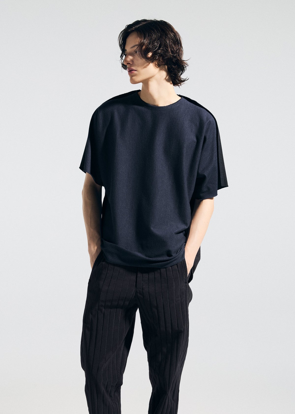 A model with medium-length wavy hair wears the TYPE-J 001 dark short-sleeved top and pinstriped pants by A-POC ABLE ISSEY MIYAKE, standing hands in pockets and gazing to the side against a plain background. view 10 of 11