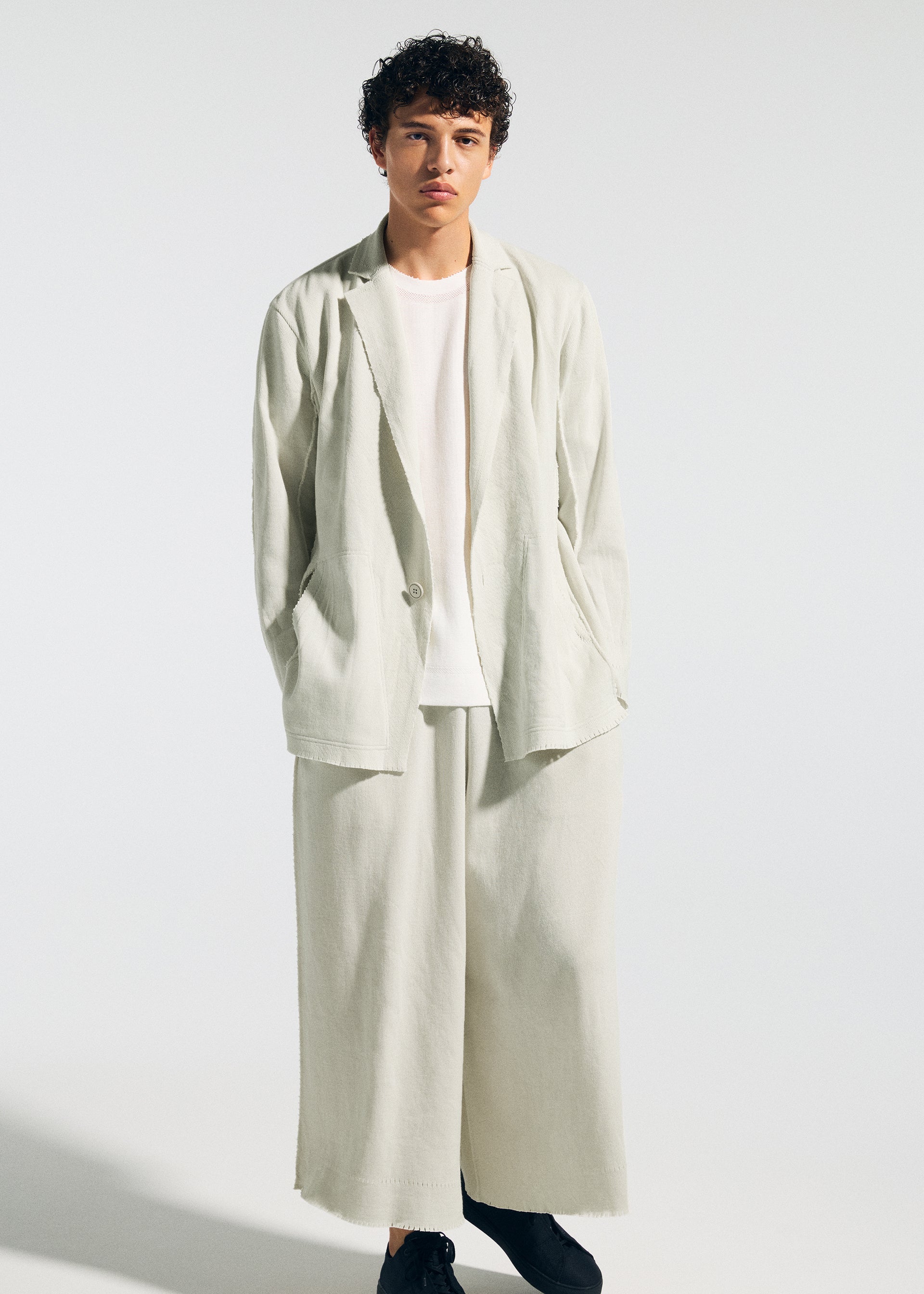 Zoomed view Against a plain background, a young person wears TYPE-A 003 by A-POC ABLE ISSEY MIYAKE: an A-POC inspired loose light suit with wide-leg pants, matching blazer, white t-shirt, and black shoes.