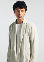 A young man with short, curly dark hair wears the TYPE-A 003 knit jacket by A-POC ABLE ISSEY MIYAKE over a white textured shirt, standing against a plain light background and gazing neutrally at the camera.