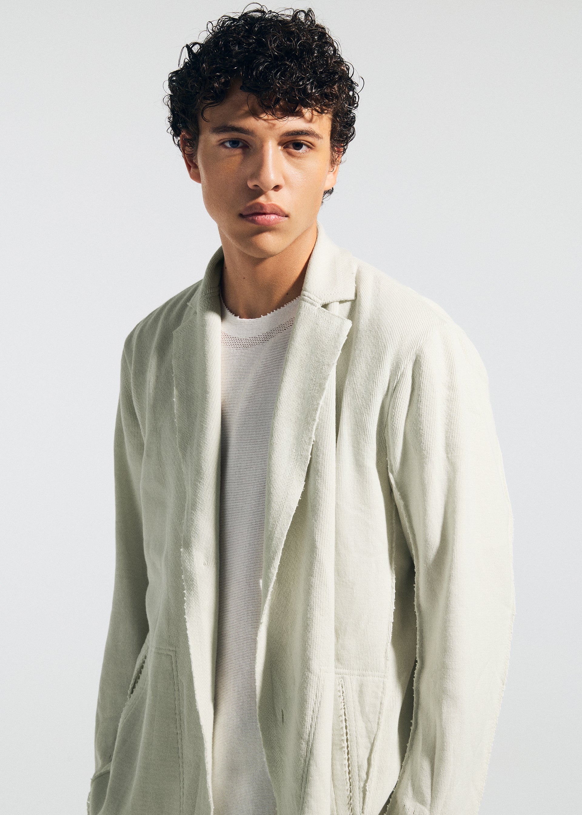 Zoomed view A young man with short, curly dark hair wears the TYPE-A 003 knit jacket by A-POC ABLE ISSEY MIYAKE over a white textured shirt, standing against a plain light background and gazing neutrally at the camera.