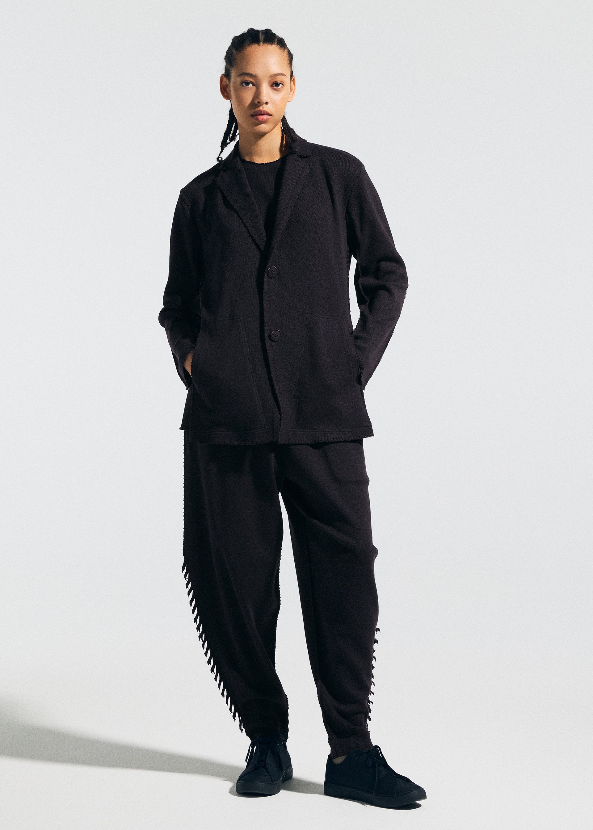 Zoomed view A person with braids faces forward, wearing the TYPE-A 003 black knit jacket and pants with ribbed side seams by A-POC ABLE ISSEY MIYAKE, paired with black sneakers against a plain background.