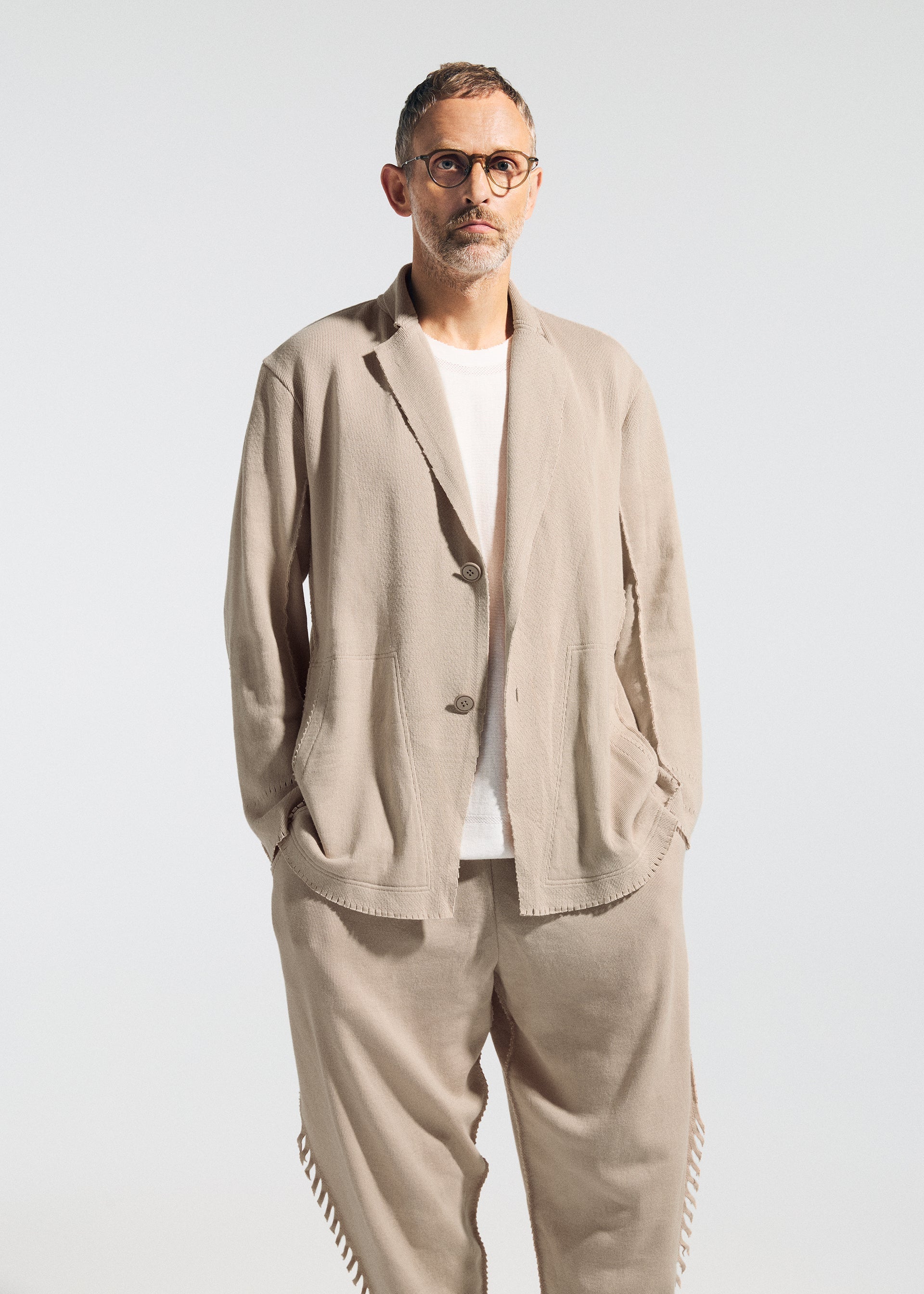 Zoomed view A man with short hair, glasses, and a trimmed beard wears the TYPE-A 003 jacket by A-POC ABLE ISSEY MIYAKE with relaxed pants and a white t-shirt. He stands hands in his pockets against a plain light background.
