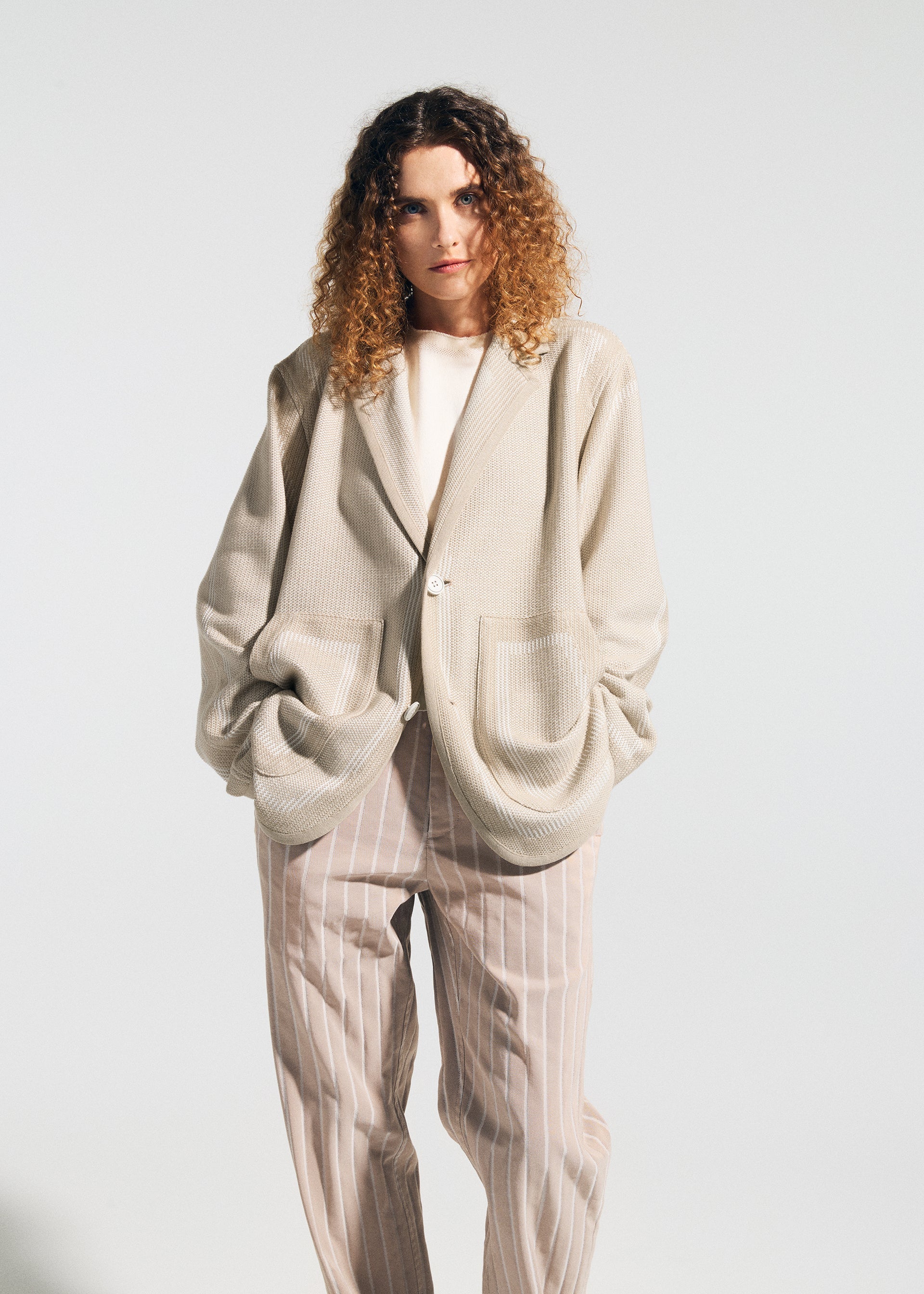 Zoomed view A woman with curly hair models the TYPE-K 008 jacket by A-POC ABLE ISSEY MIYAKE, in beige with large pockets, over a white top and beige pinstriped trousers against a plain light background.