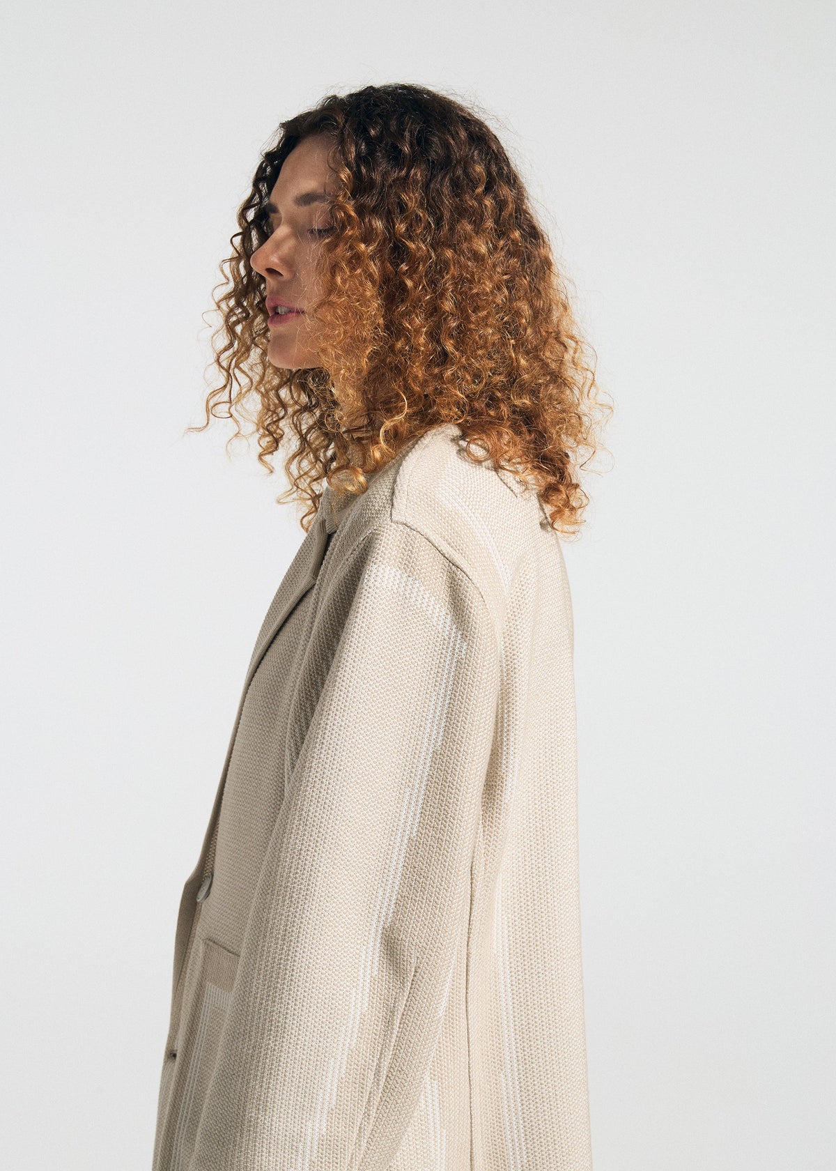 A person with shoulder-length curly hair models the TYPE-K 008 textured knit jacket by A-POC ABLE ISSEY MIYAKE in light beige, standing in profile against a plain white background. view 7 of 11