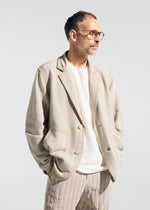 A man with short hair, beard, and glasses models the TYPE-K 008 blazer by A-POC ABLE ISSEY MIYAKE—light beige, wrinkle-resistant—over a white shirt and beige pinstriped pants, standing hands-in-pockets against a plain background.