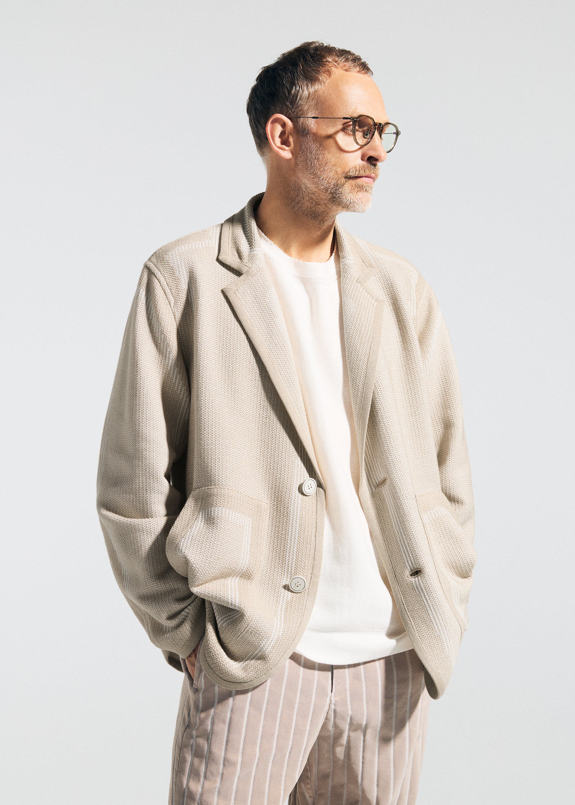 Zoomed view A man with short hair, beard, and glasses models the TYPE-K 008 blazer by A-POC ABLE ISSEY MIYAKE—light beige, wrinkle-resistant—over a white shirt and beige pinstriped pants, standing hands-in-pockets against a plain background.